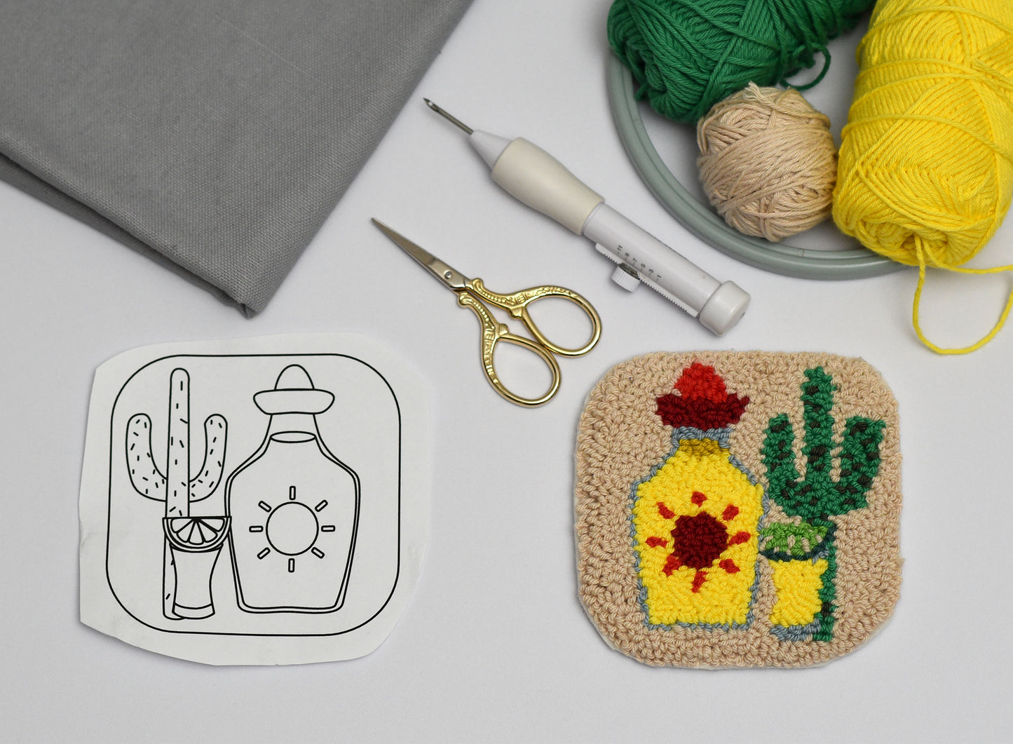 Embroidery hoop with colorful embroidery cactus and Tequila bottle design, yarn, scissors, and fabric on a light gray background