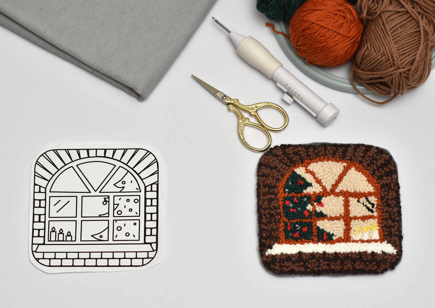 Punch needled coaster with house design next to yarn, crochet hook, and scissors on a white background