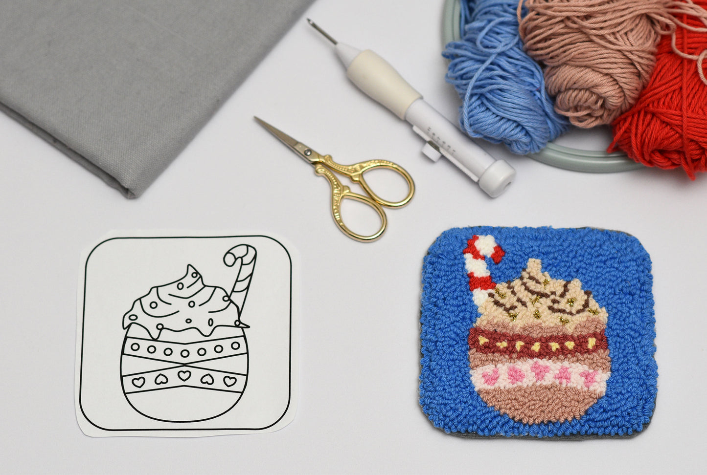 Embroidery hoop with a completed punch needle coaster design of a hot cocoa mug, next to a template and embroidery supplies on a white surface.