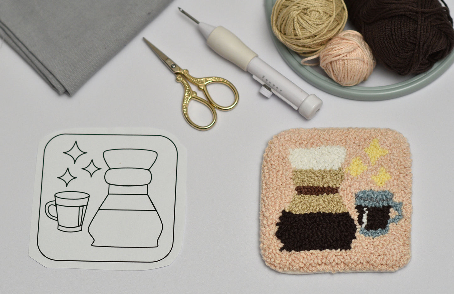 Handmade punch needle coaster with coffee-themed design including chemex next to a black and white outline drawing on a light gray surface.