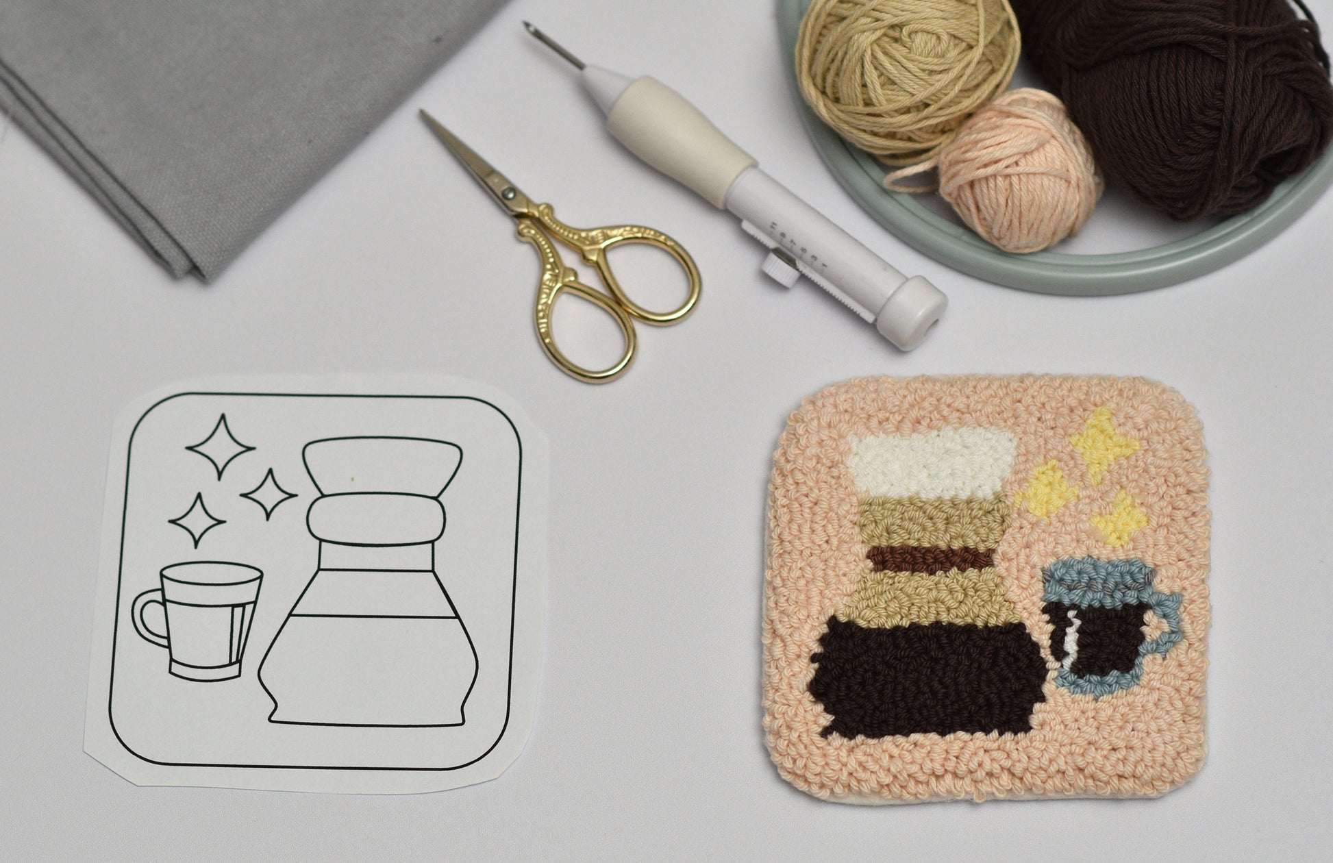 Handmade punch needle coaster with coffee-themed design including chemex next to a black and white outline drawing on a light gray surface.