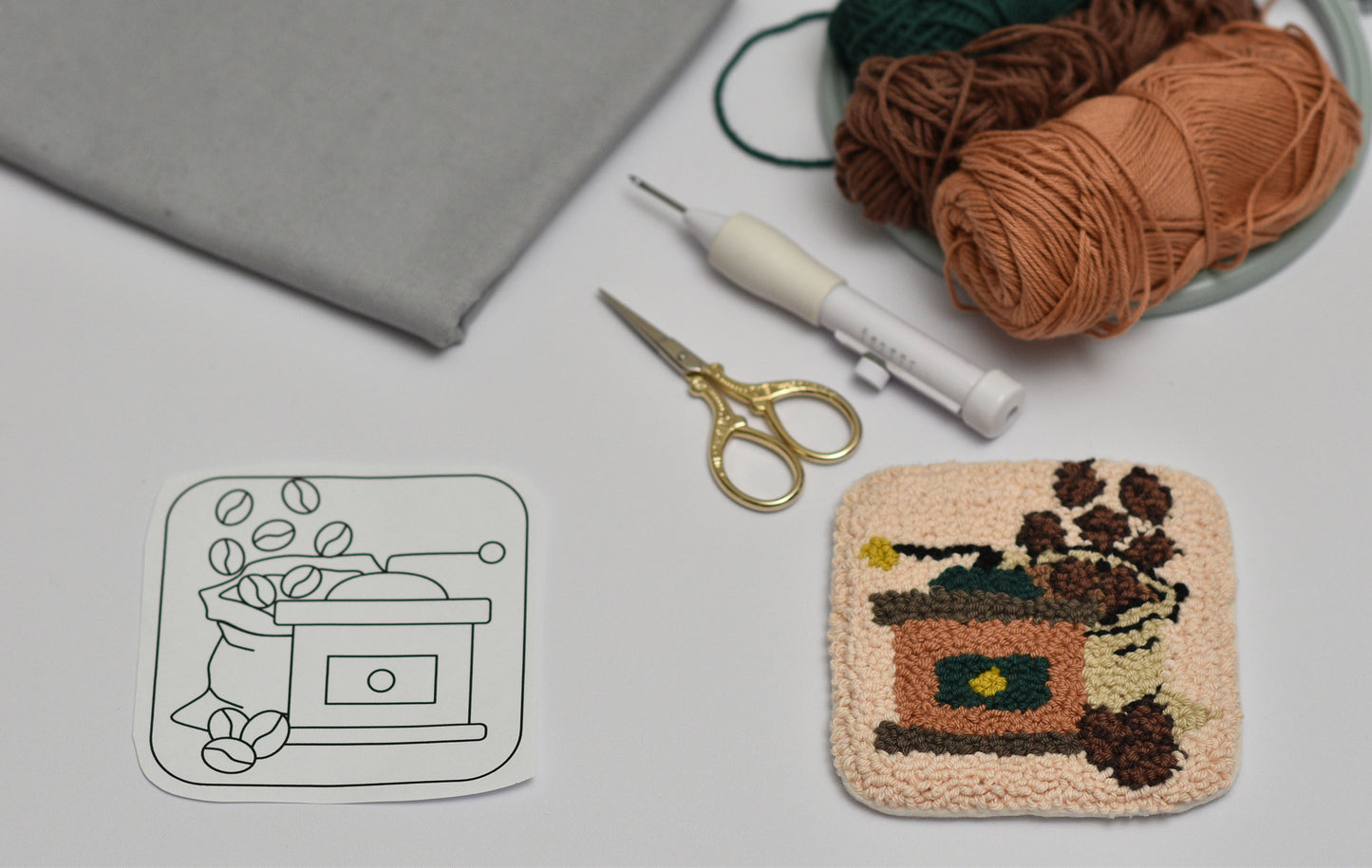 Handmade punch needle coaster project with yarn, scissors, and design sketch of a coffee grinder on a gray surface