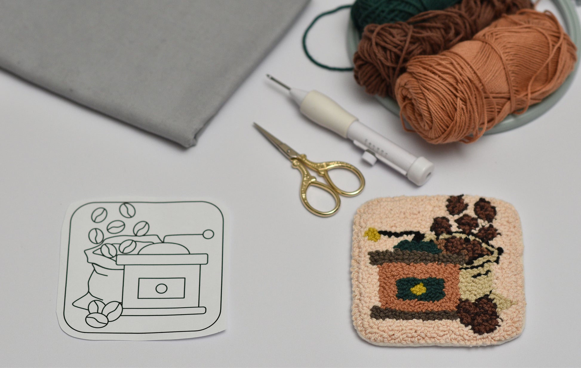 Handmade punch needle coaster project with yarn, scissors, and design sketch of a coffee grinder on a gray surface