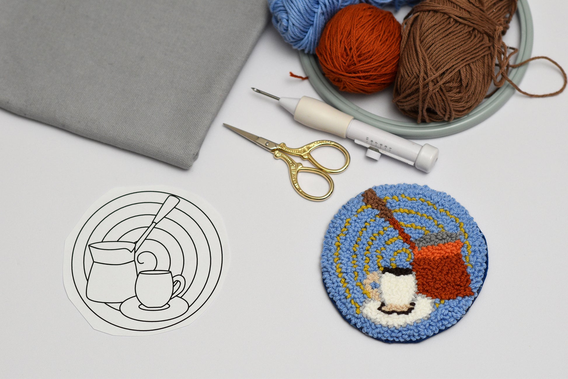 Embroidery hoop with a completed embroidery coaster design of a Turkish coffee pot and cup, next to yarn and embroidery tools on a light gray background.