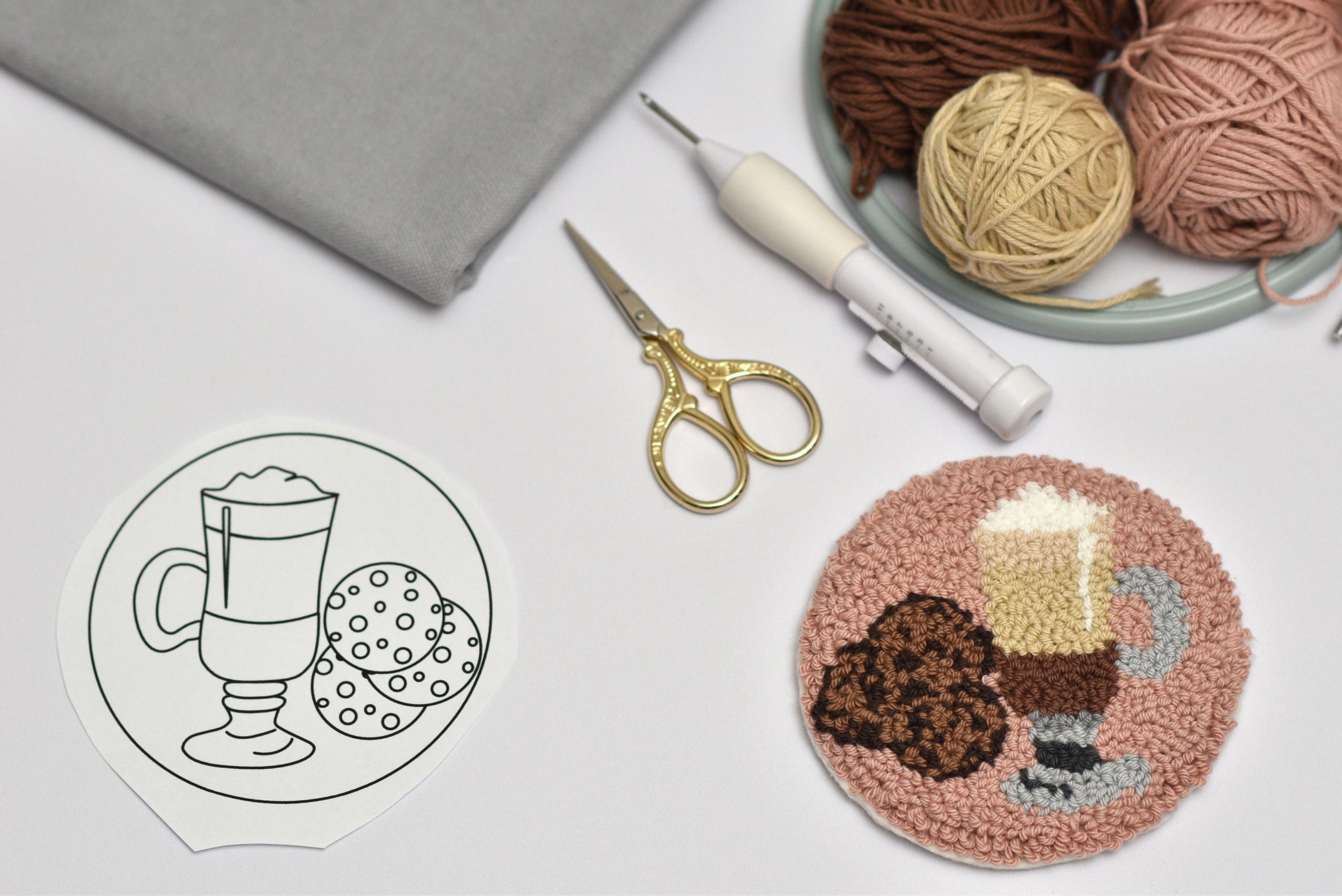 Punch needle coaster with coffee design, yarn balls, scissors, and punch needle hook on a white surface.