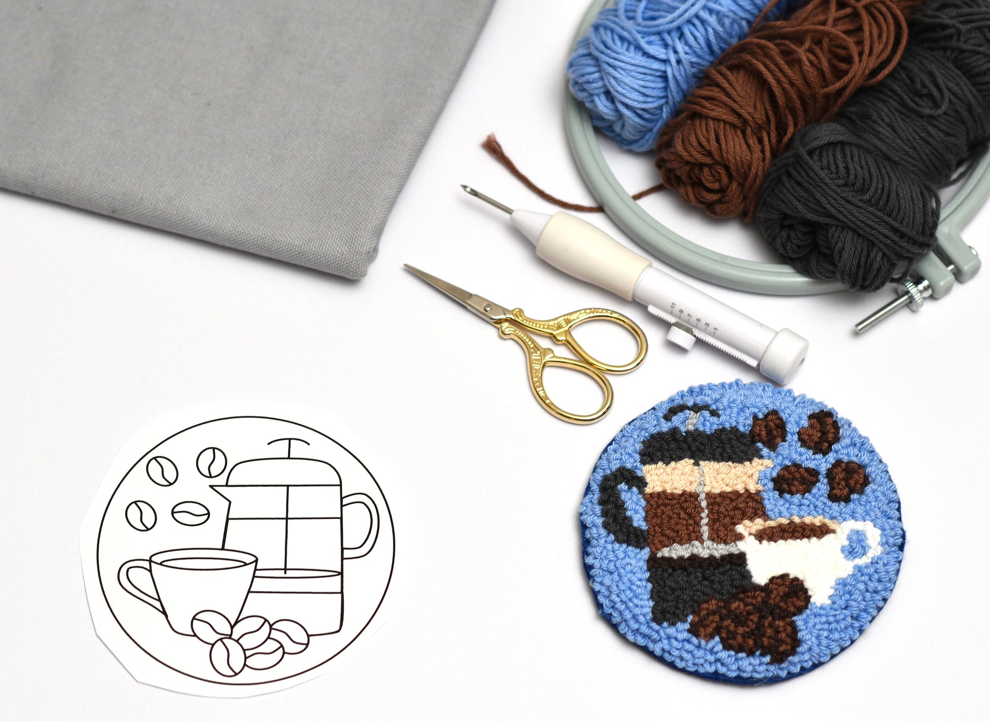 Punch needled coaster with coffee-themed design, yarn, scissors, and fabric on a white surface.