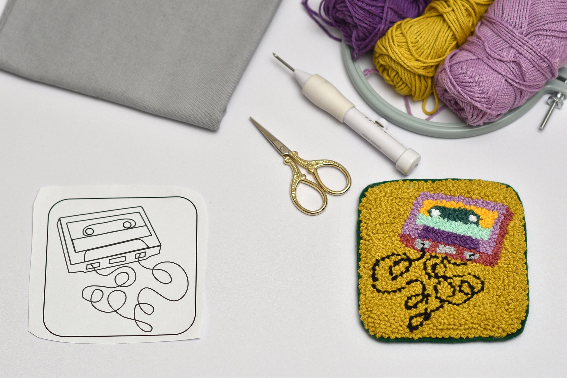 Embroidered coaster with a cassette tape design, yarn, scissors, and crochet hook on a white surface.