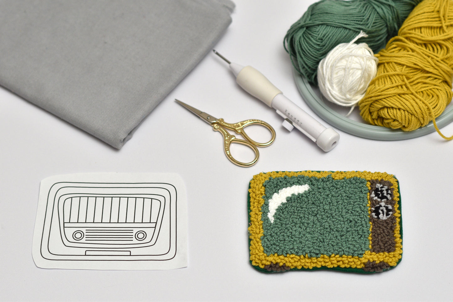 Punch needle coaster with retro TV design, yarn, scissors, and other crafting tools on a white surface