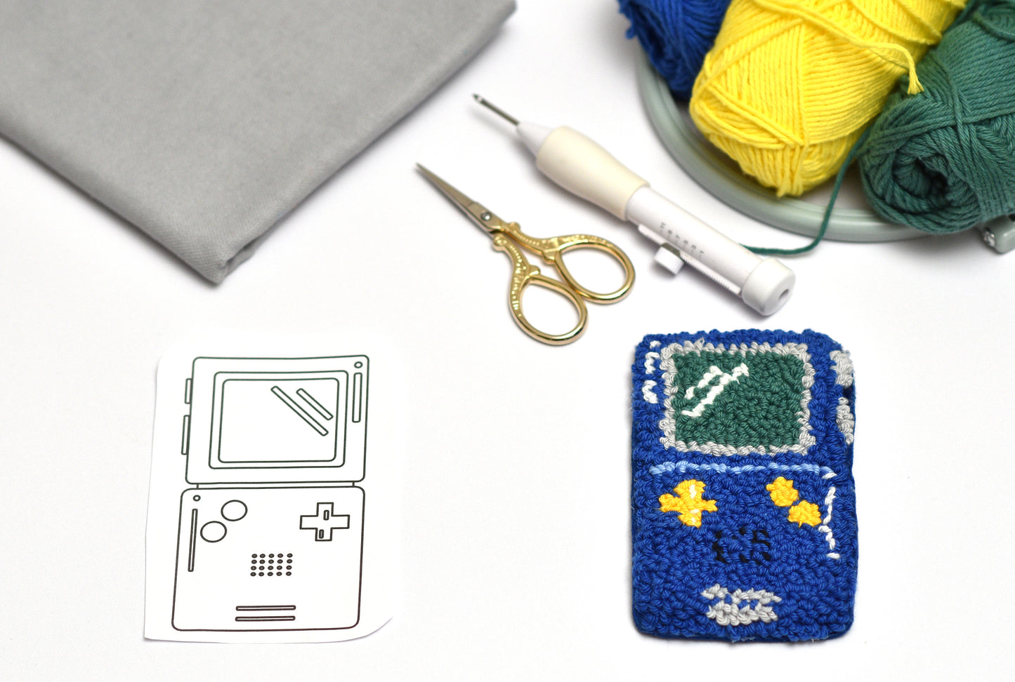 Embroidered Game Boy patterned coaster with yarn, scissors, and a template on a white surface