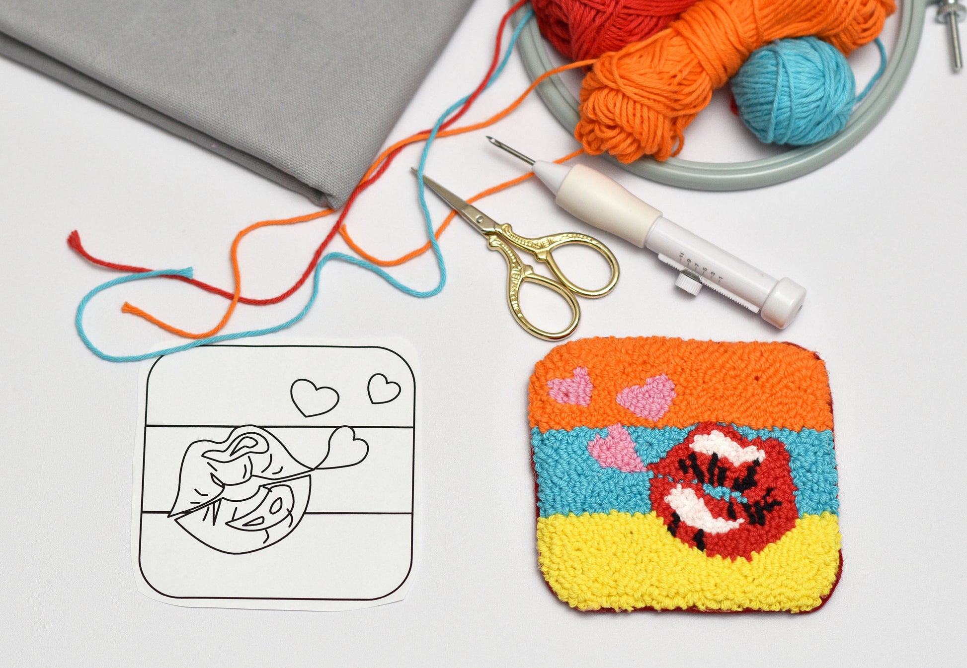 Embroidered coaster with colorful design next to yarn, punch needle hook, and scissors on a white surface.