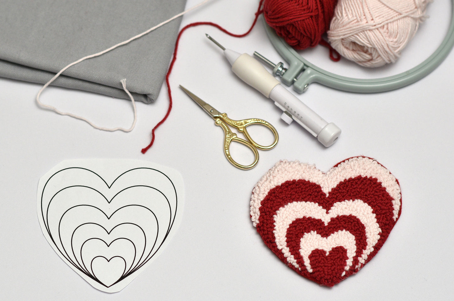 Crocheted heart pattern next to yarn, scissors, and an embroidery hoop on a light gray background