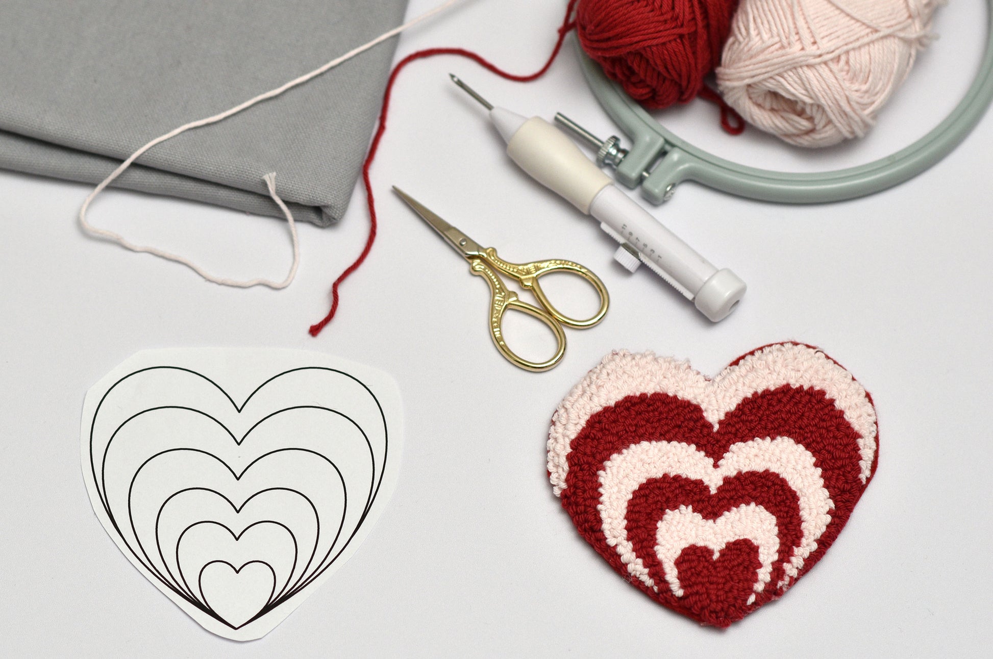 Crocheted heart pattern next to yarn, scissors, and an embroidery hoop on a light gray background