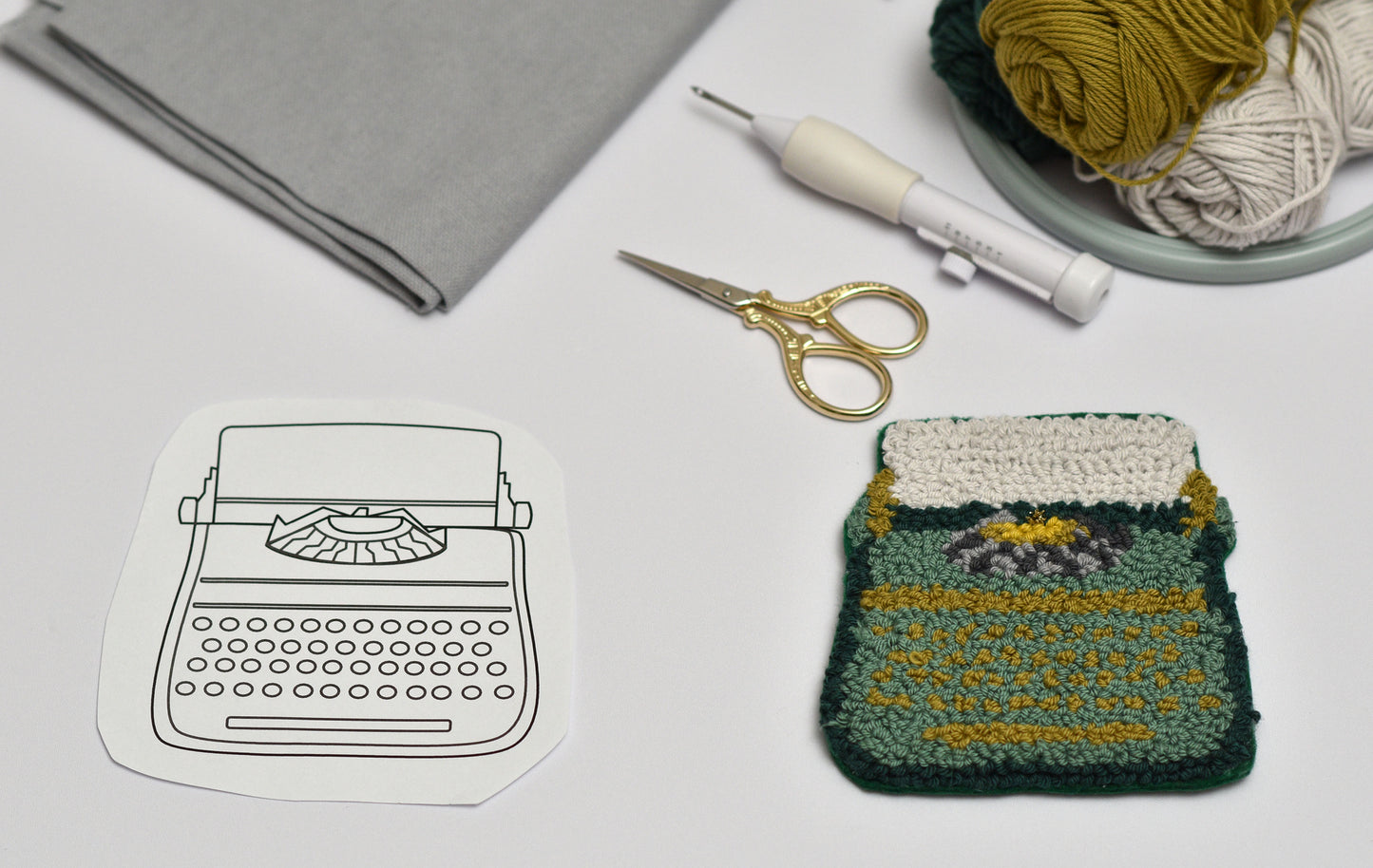 Embroidered typewriter coaster next to a paper cut out of a typewriter on a table with yarn and scissors.