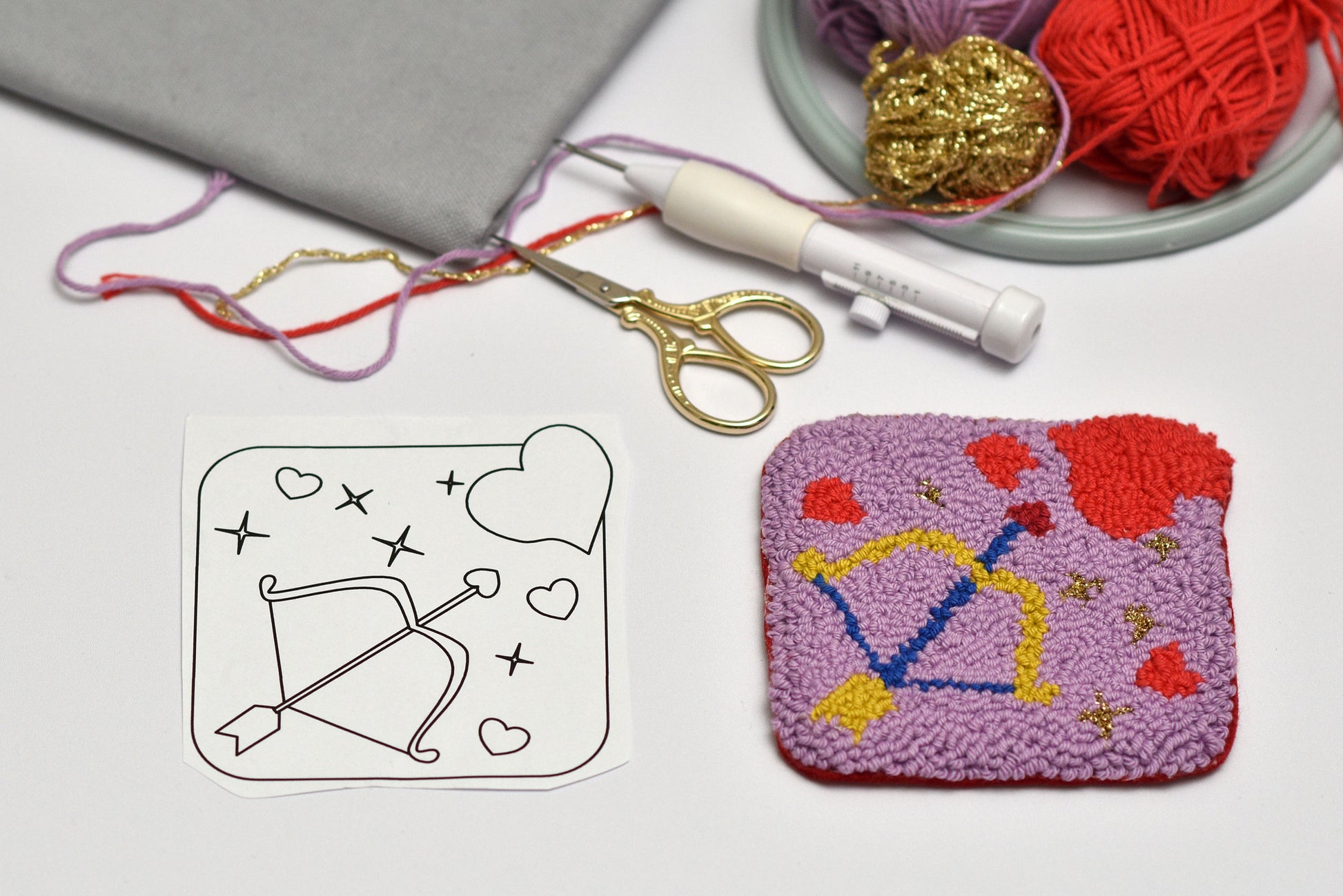 Embroidery project with a pattern, scissors, thread, and completed embroidered square coaster on a gray surface.