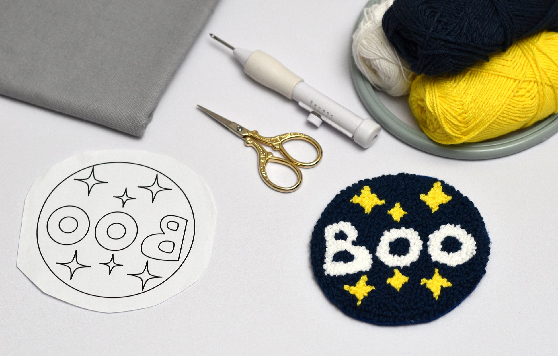 Punch needled Coaster with 'BOO' text and star patterns next to a white template, scissors, and yarn on a light gray background.