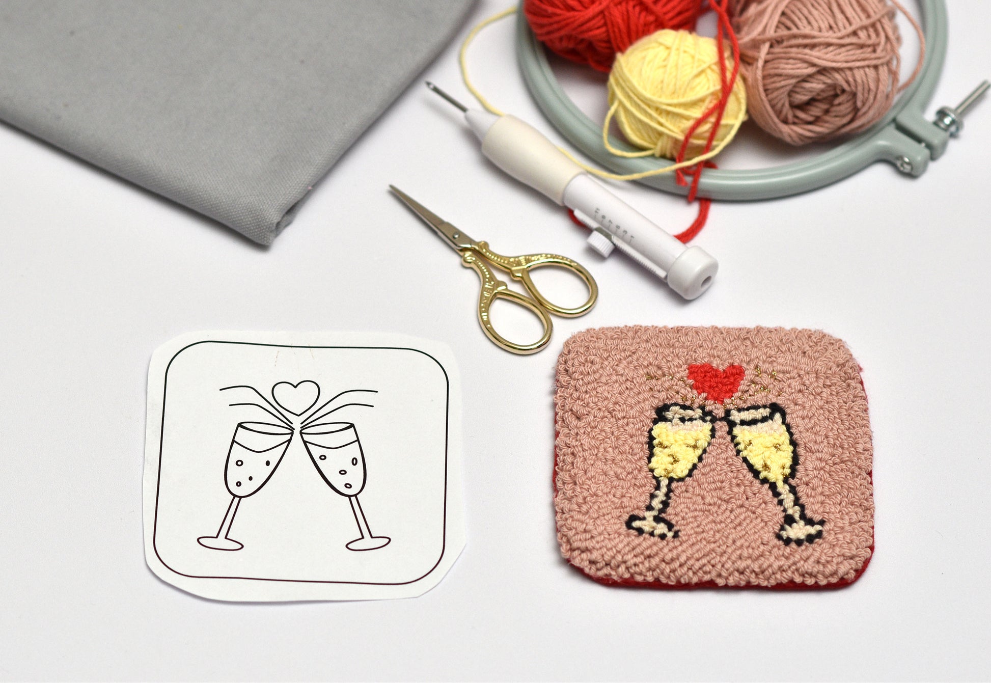Embroidery hoop with yarn, scissors, and a completed embroidered coaster with champagne design on a white background