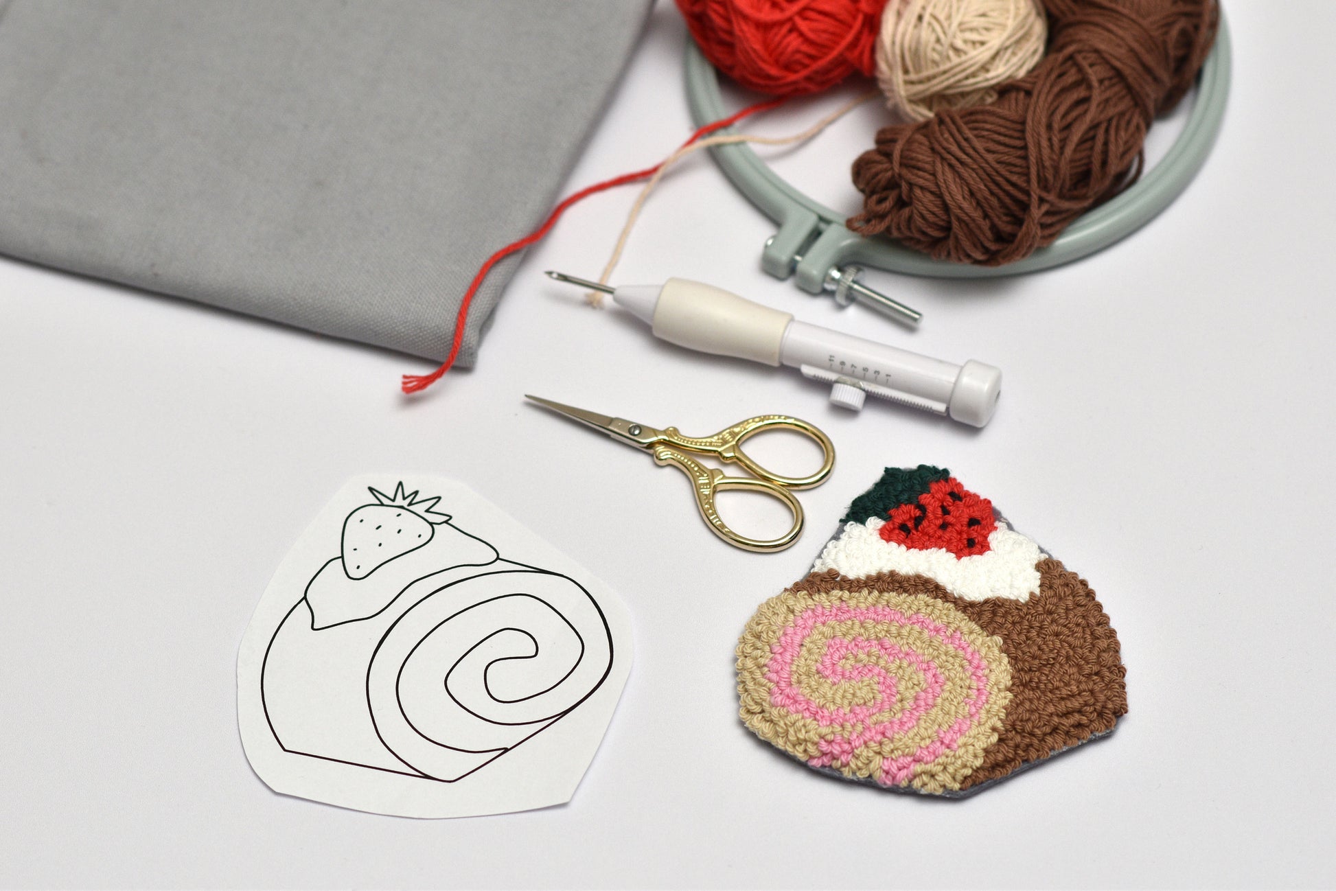 Embroidery hoop, yarn, scissors, and embroidered fabric with a cake designed coaster on a light gray surface