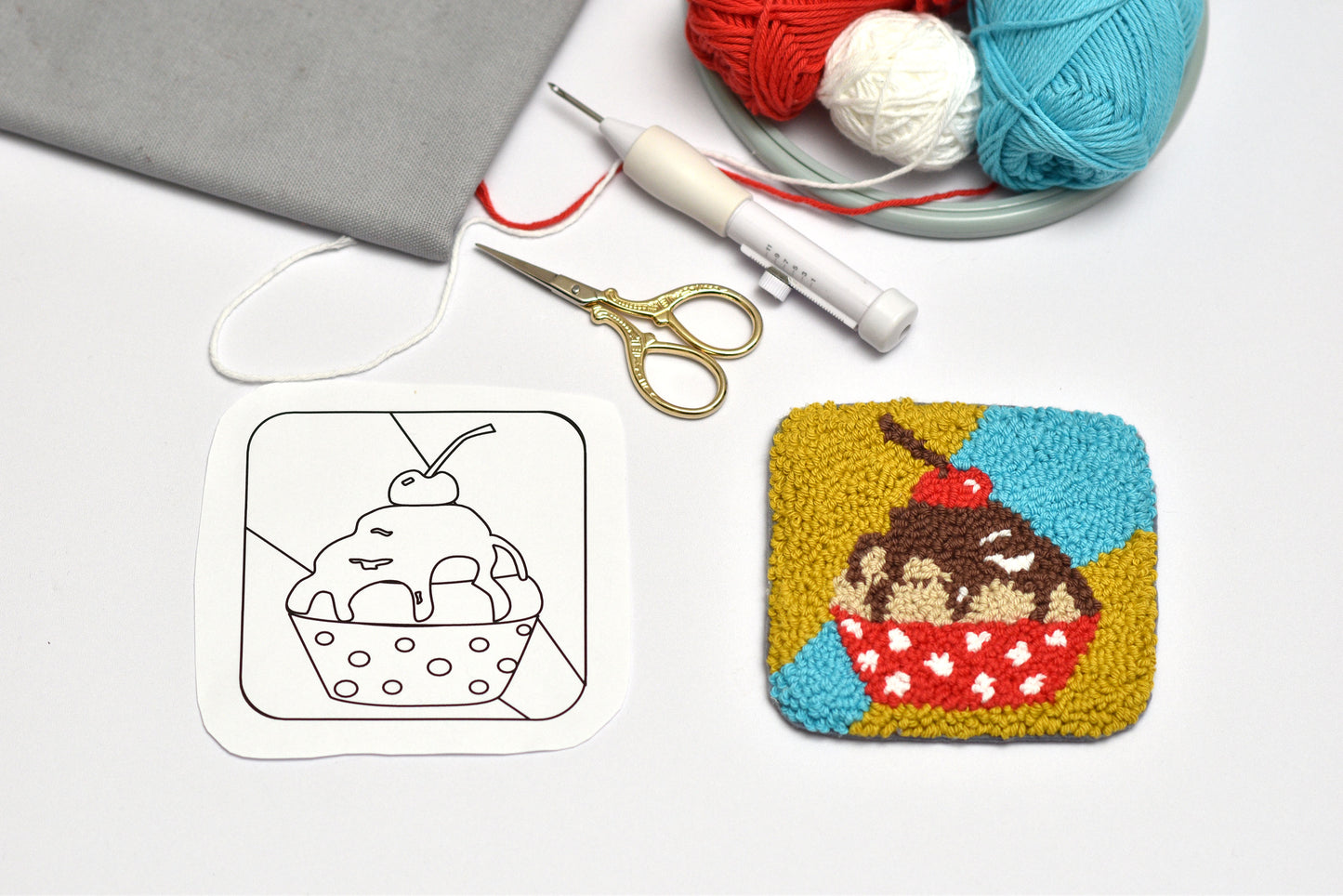 Completed and outlined embroidered coaster with cupcake design with yarn and tools on a white surface