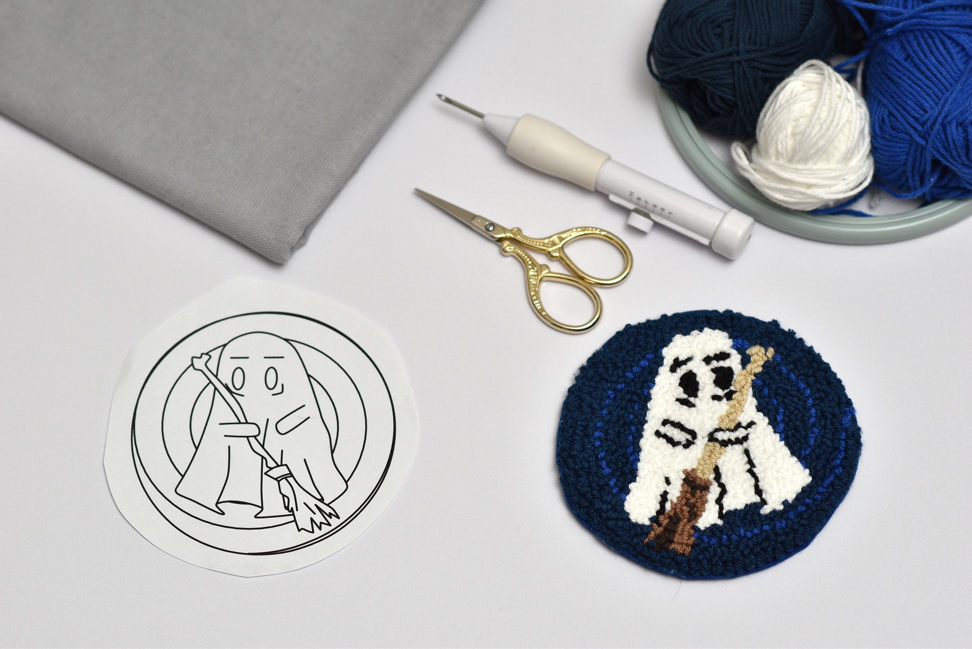Embroidery hoop with a completed coaster design of a ghost character holding a broom, next to a black and white outline drawing on fabric.