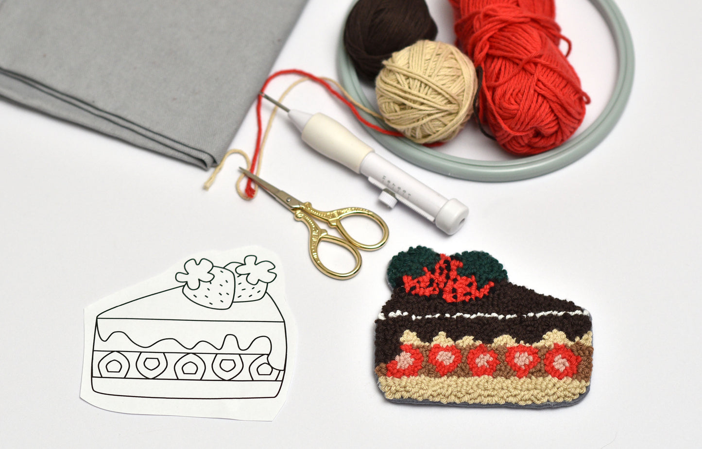 Embroidered cake-shaped item next to a coloring book page with a cake illustration, yarn, and scissors on a white surface.