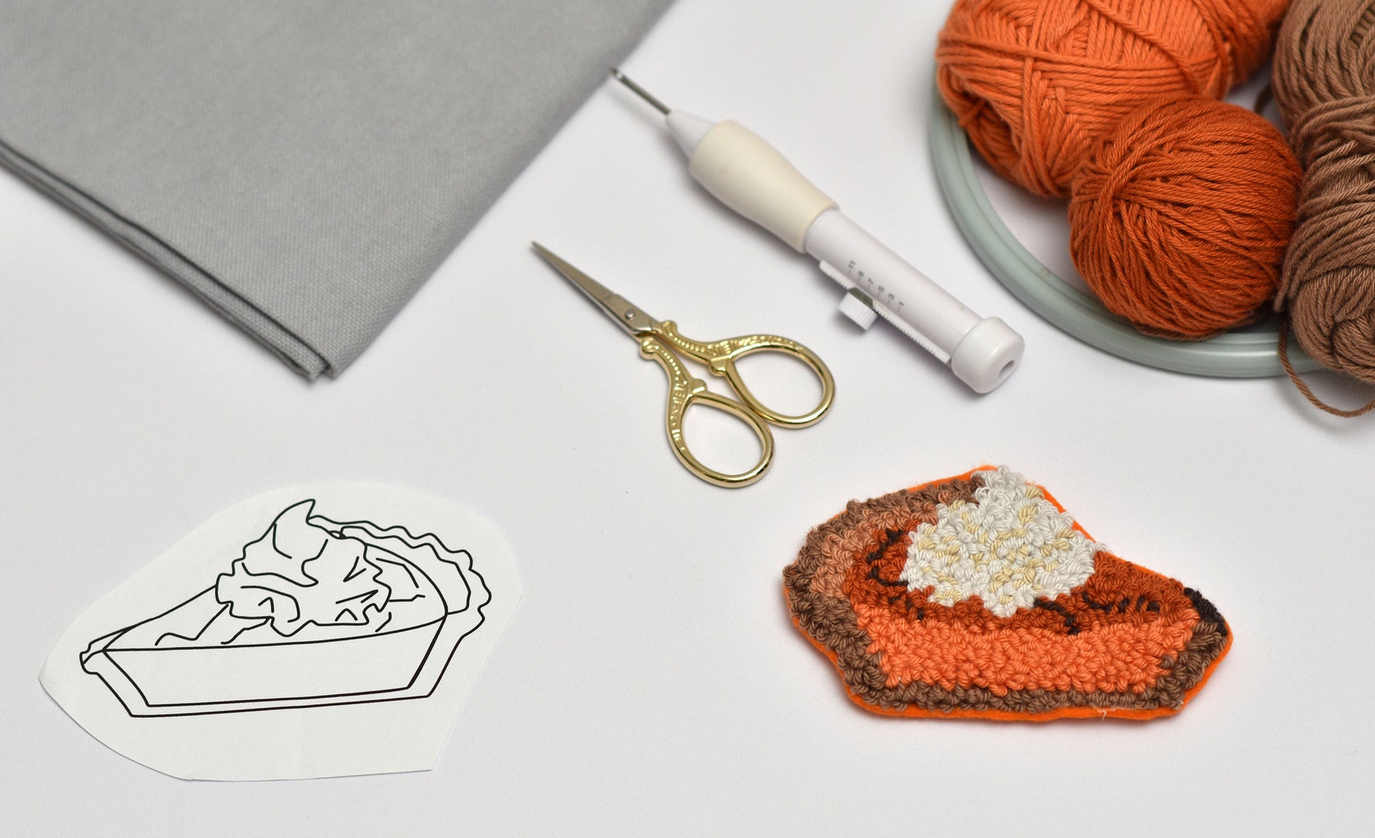 Punch needled coaster with pumpkin pie slice design with yarn, scissors, and a template on a table.