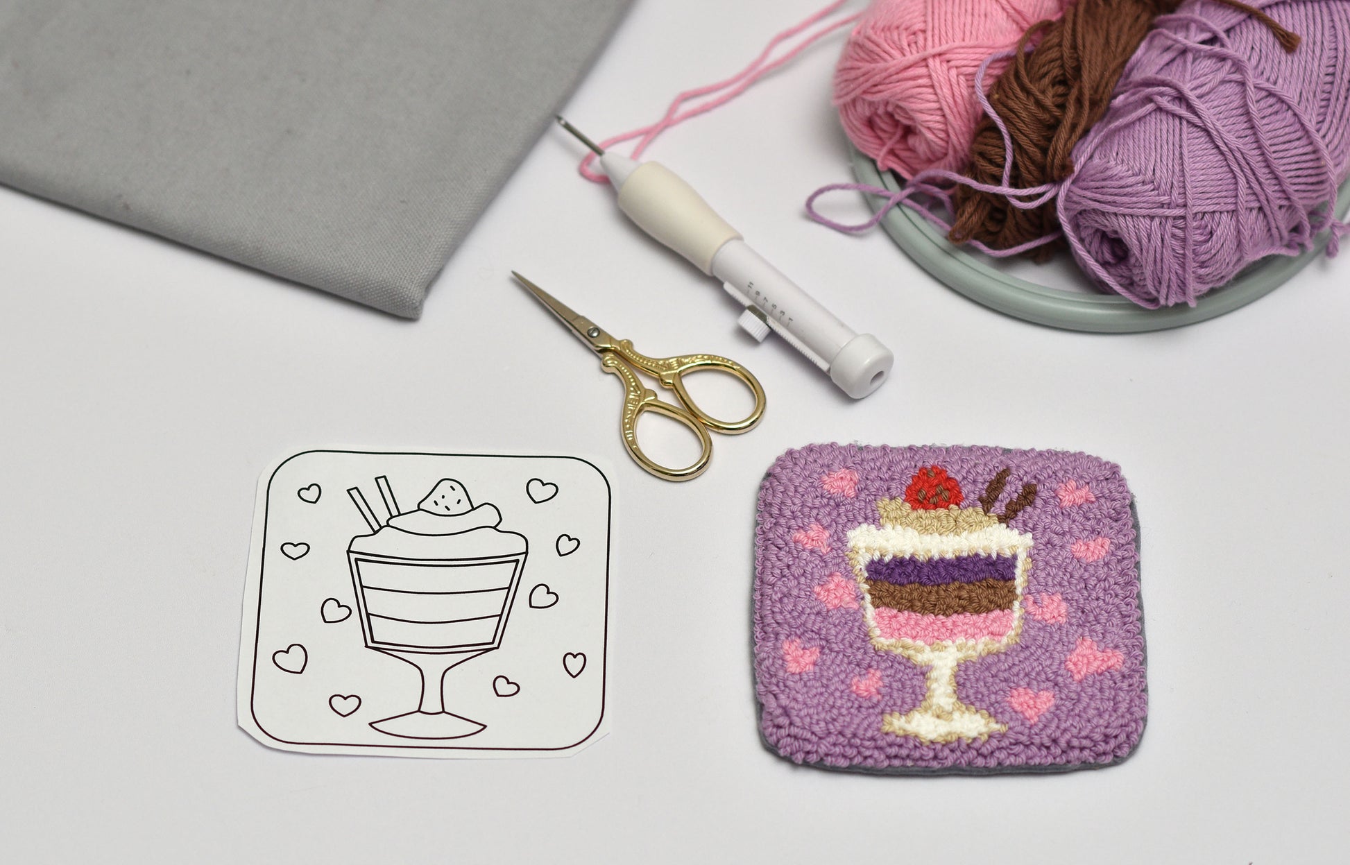 Embroidered coaster with parfait dessert design next to a paper template on a table with yarn and scissors.