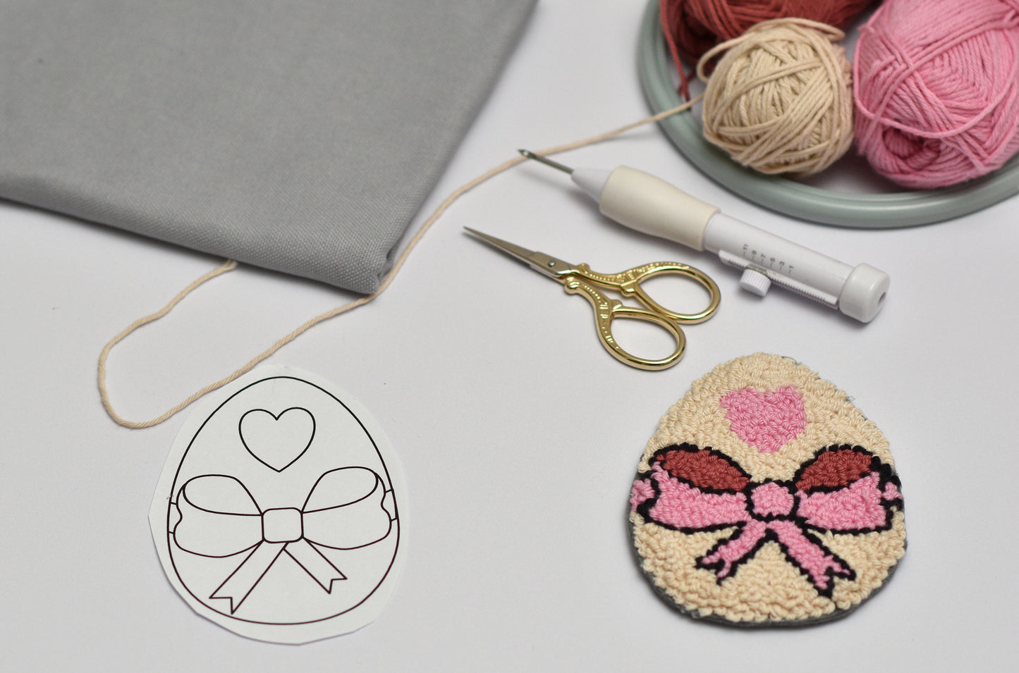 Embroidery hoop with a completed Easter Egg coaster with pink ribbon, scissors, and yarn on a gray surface.