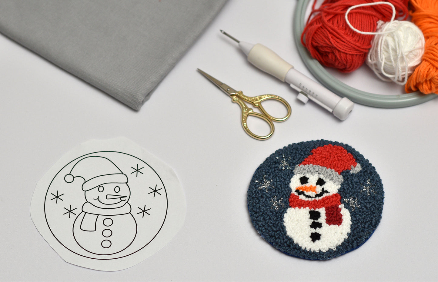 Punch needled snowman coaster and outline on fabric with yarn, scissors, and punch needle hook.