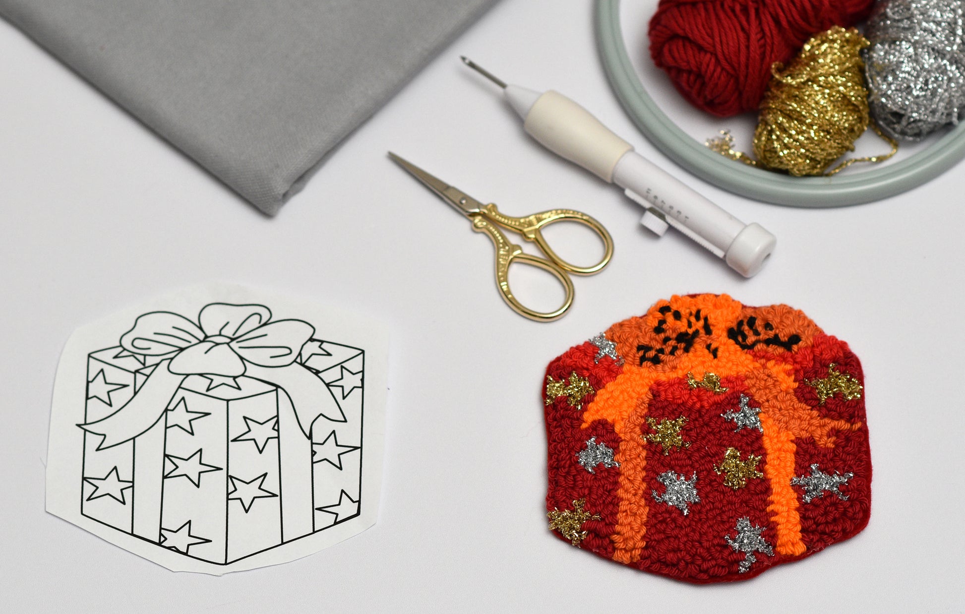 Gift box designed punch needle coaster with decorative elements next to a black and white outline, scissors, and yarn on a light gray background.