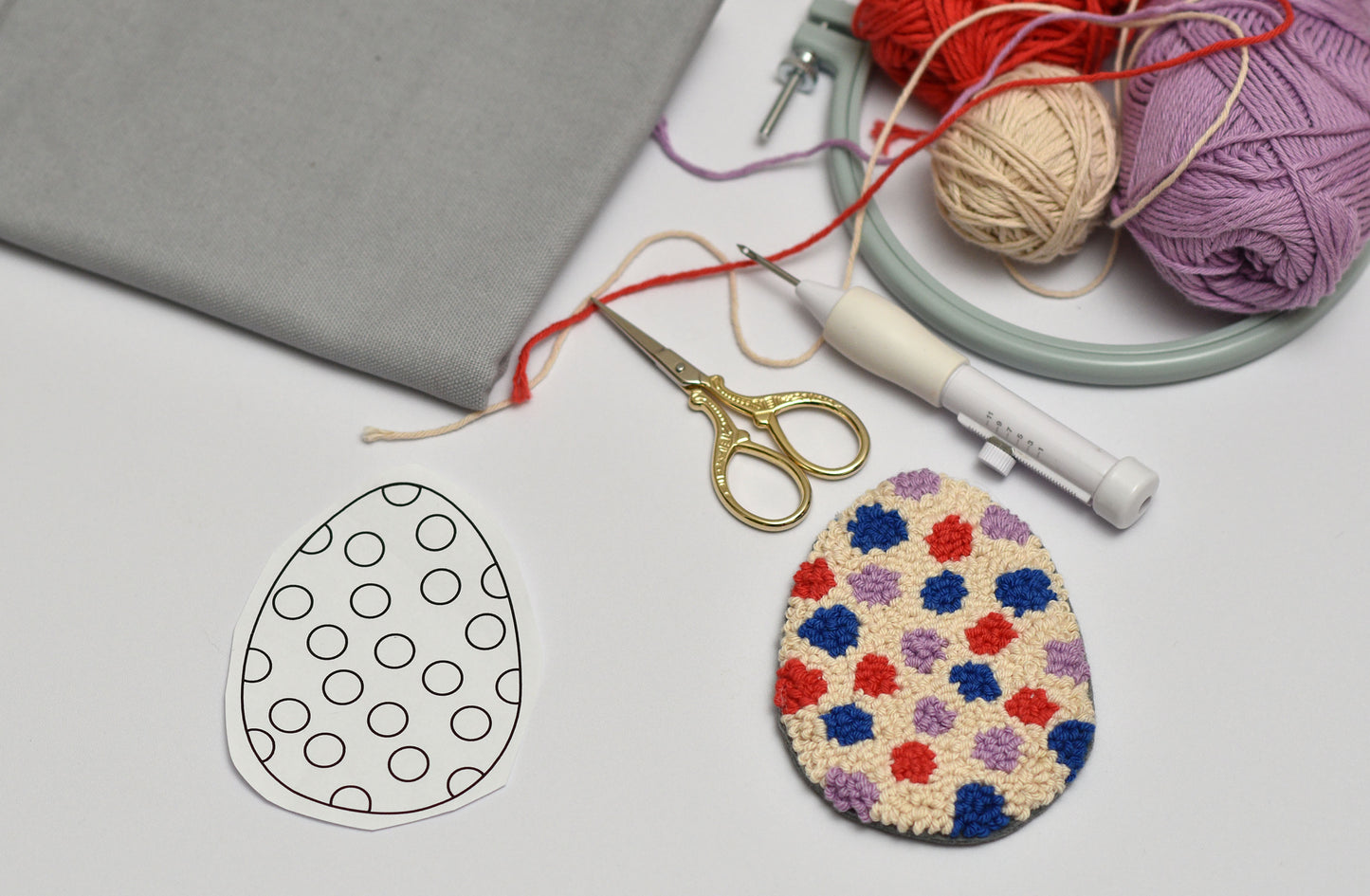 Punch needled Easter egg, yarn, scissors, and a paper template on a gray surface