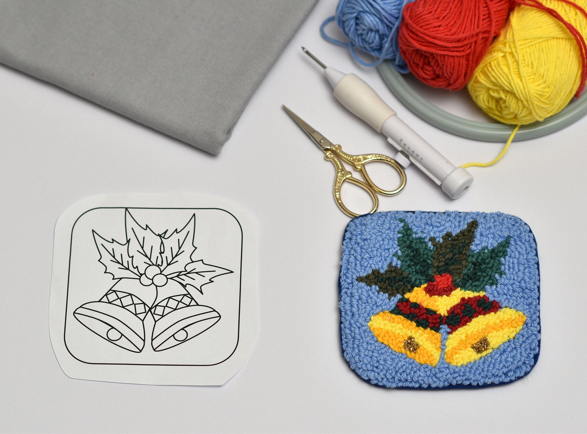 Christmas themed coaster with bell design next to a line drawing, yarn, and crochet tools on a gray surface.