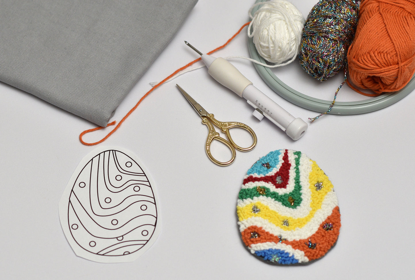 Colorful embroidered Easter egg, patterned outline, scissors, and yarn on a light surface.