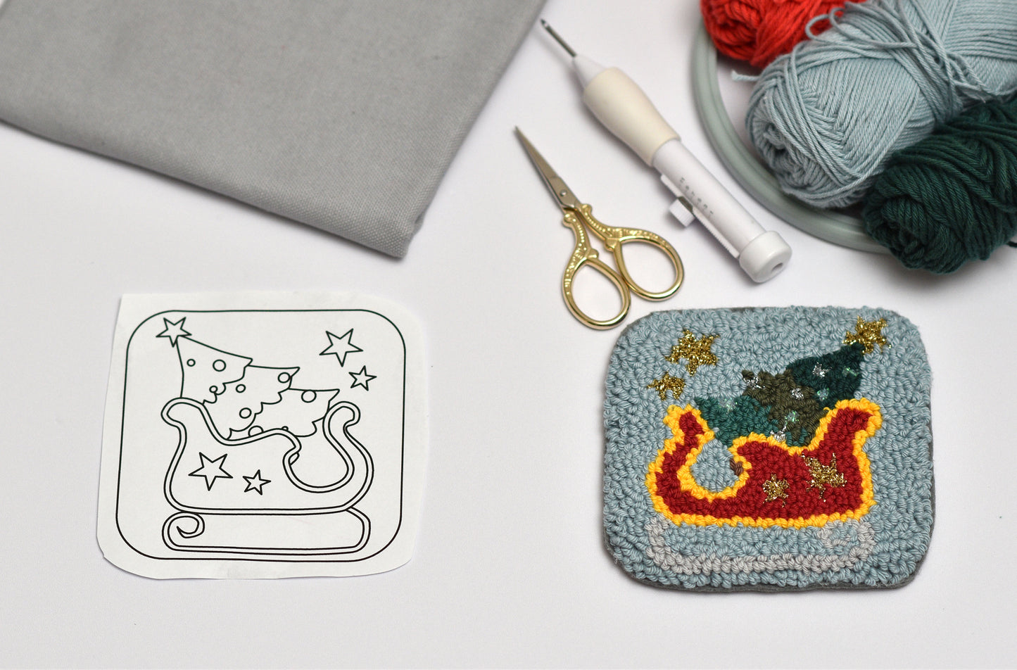 Punch needled coaster with Christmas design next to a template on a table with yarn and tools.