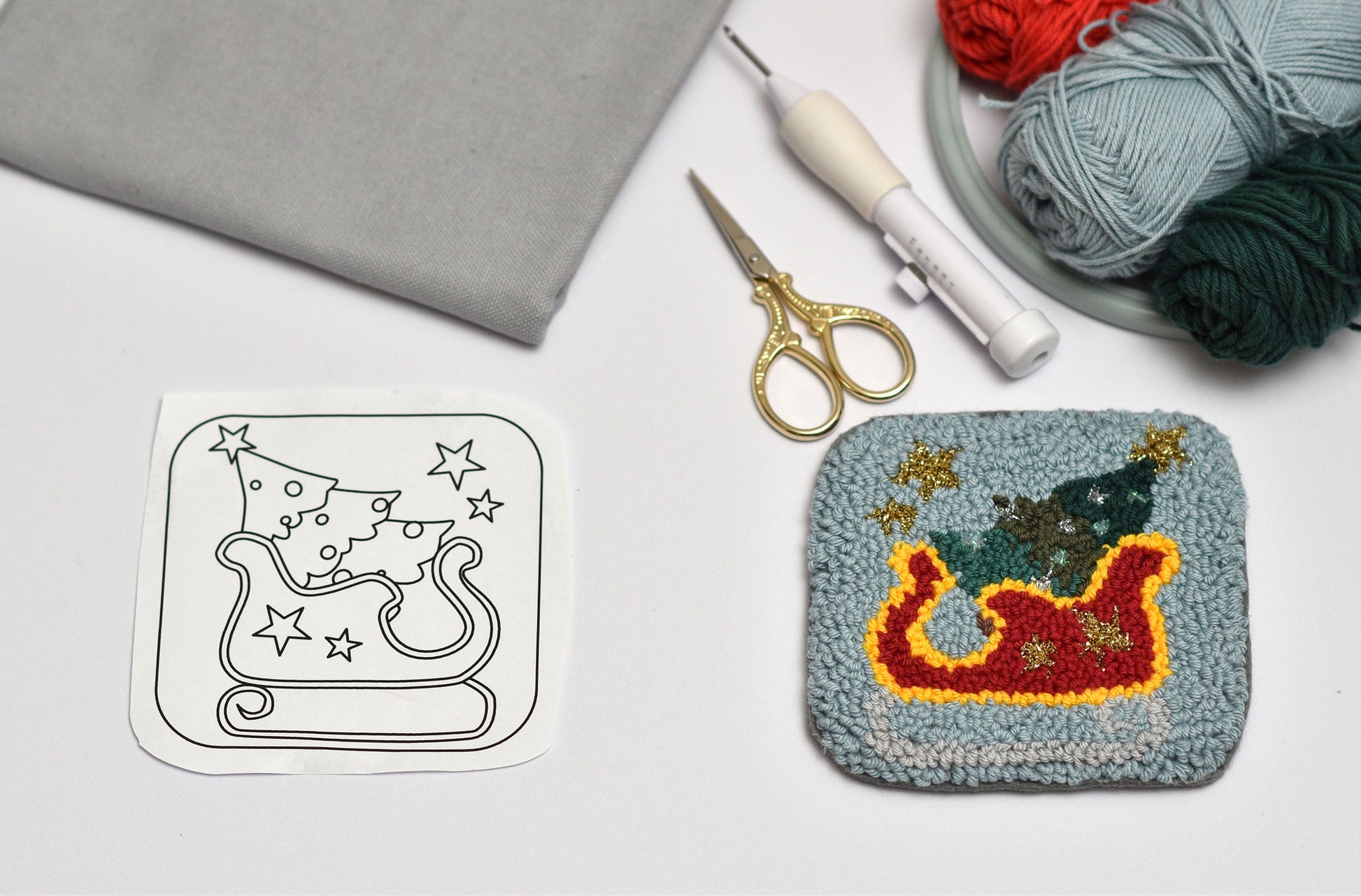 Punch needled coaster with Christmas design next to a template on a table with yarn and tools.