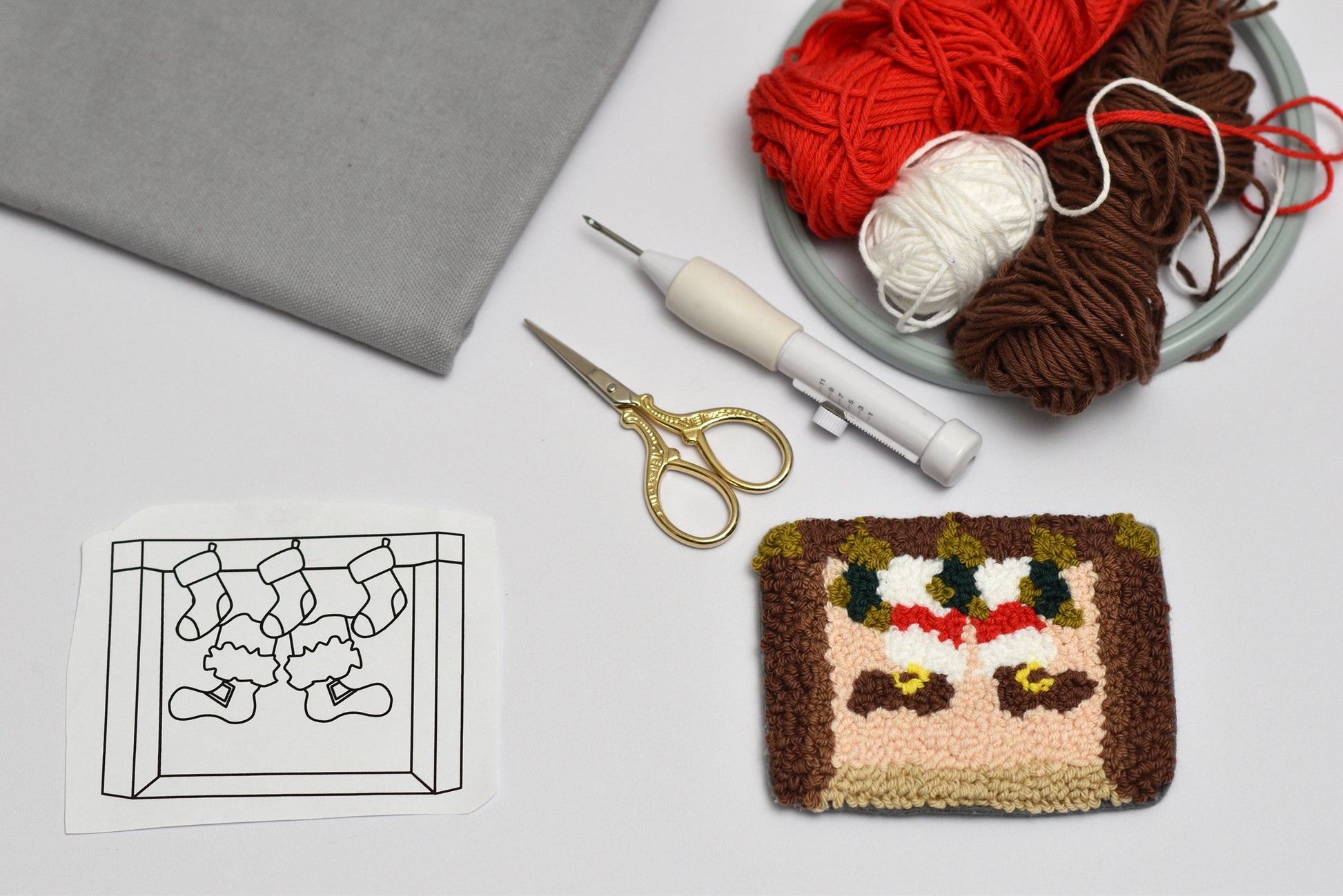 Punch needled coaster with Santa stuck in a chimney design next to yarn, scissors, and a pattern sheet on a light gray surface.