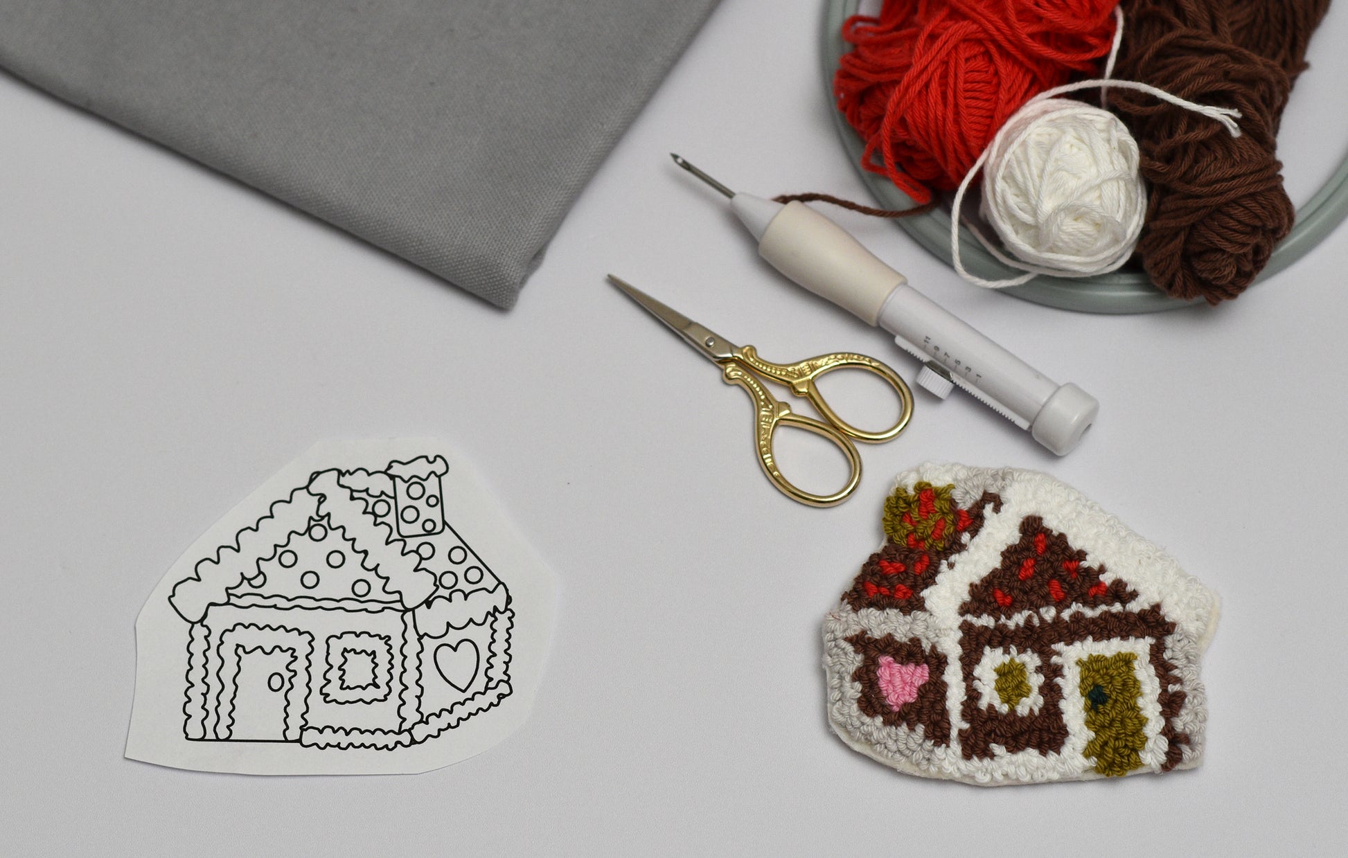 Embroidered gingerbread house pattern next to a completed punch needle gingerbread house coaster with yarn and punch needle tools on a gray background.