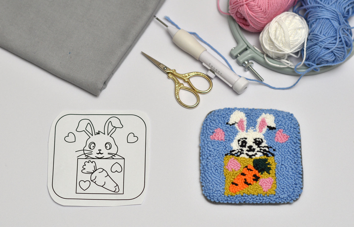 Embroidered bunny with carrot design next to a black and white outline on a gray surface with yarn and tools.