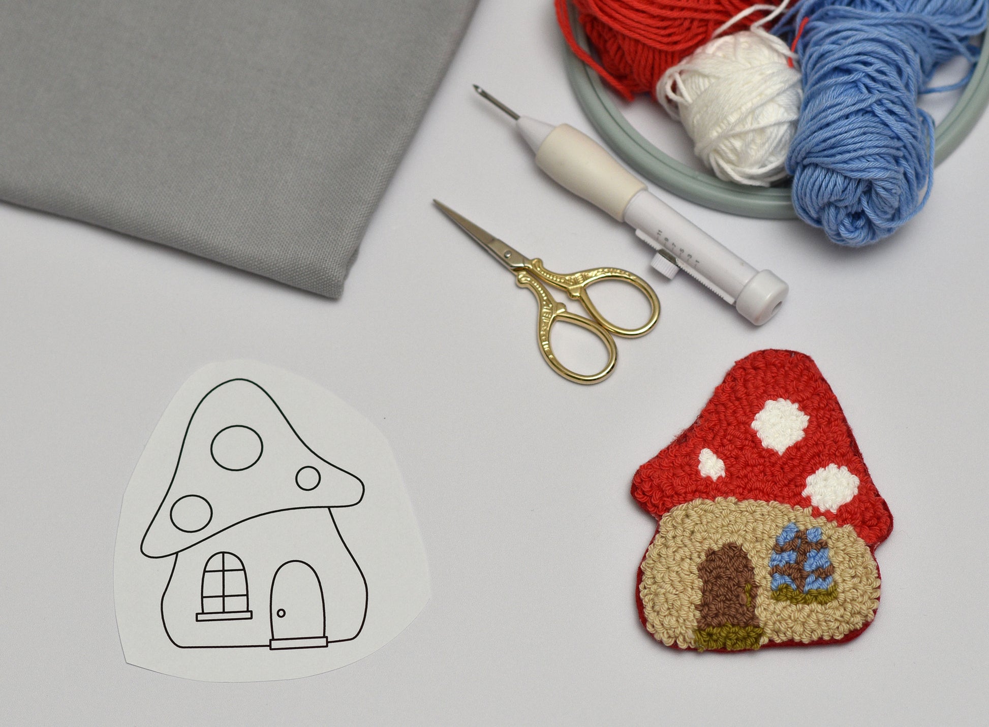 Punch needle coaster with mushroom house design with yarn, scissors, and a template on a gray surface