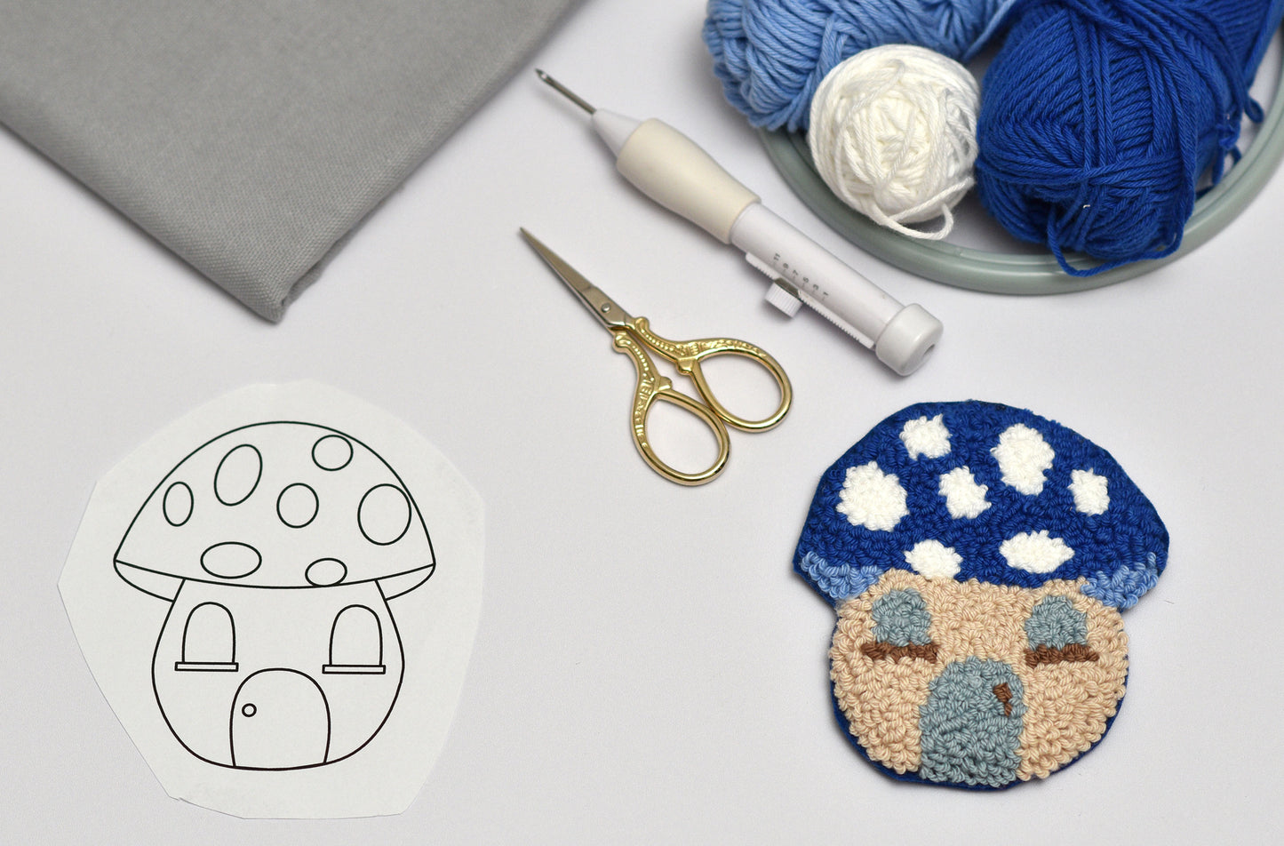 Handmade punch needled mushroom-shaped coaster with yarn, scissors, and a template on a light surface.