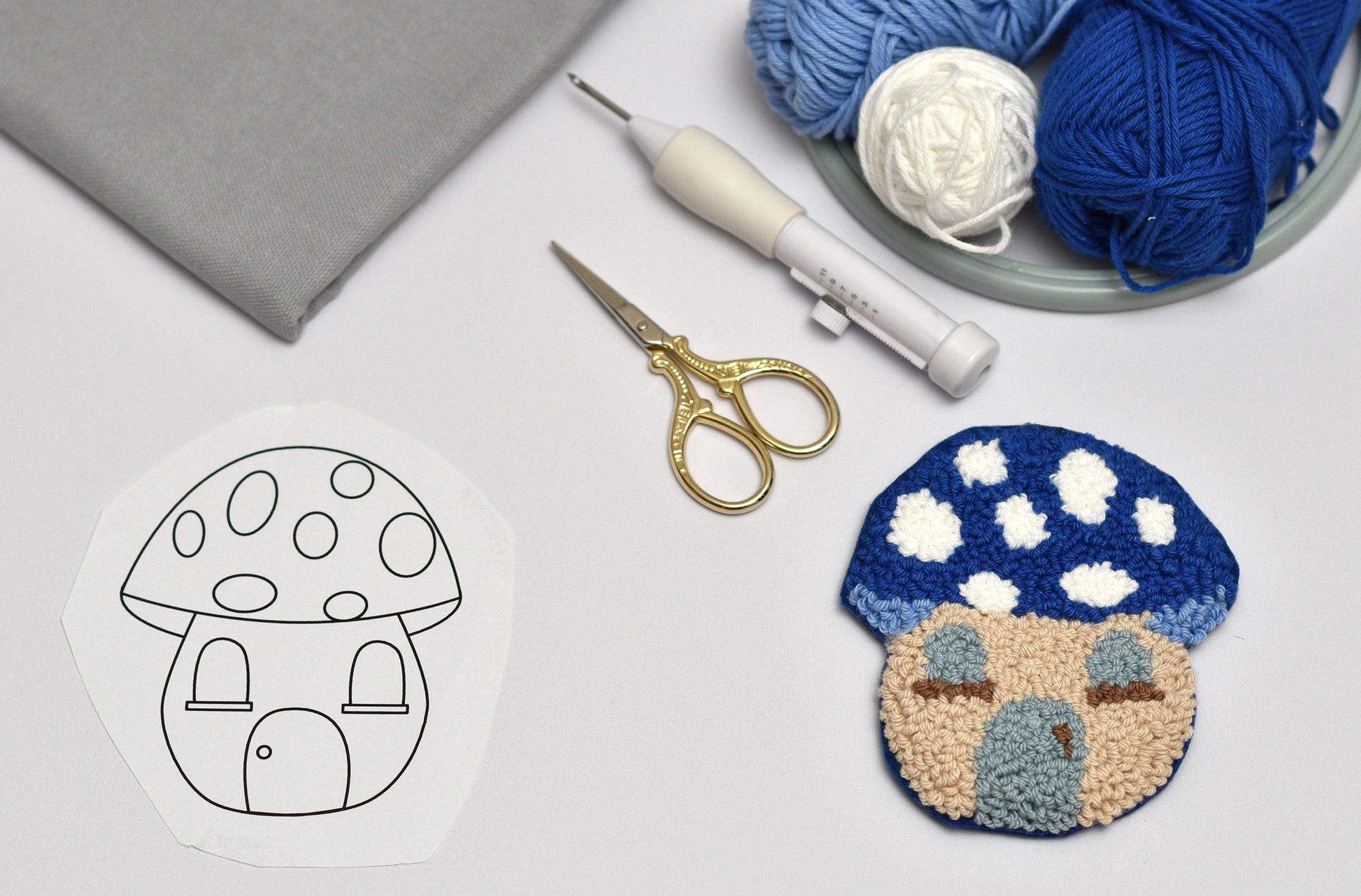 Handmade punch needled mushroom-shaped coaster with yarn, scissors, and a template on a light surface.
