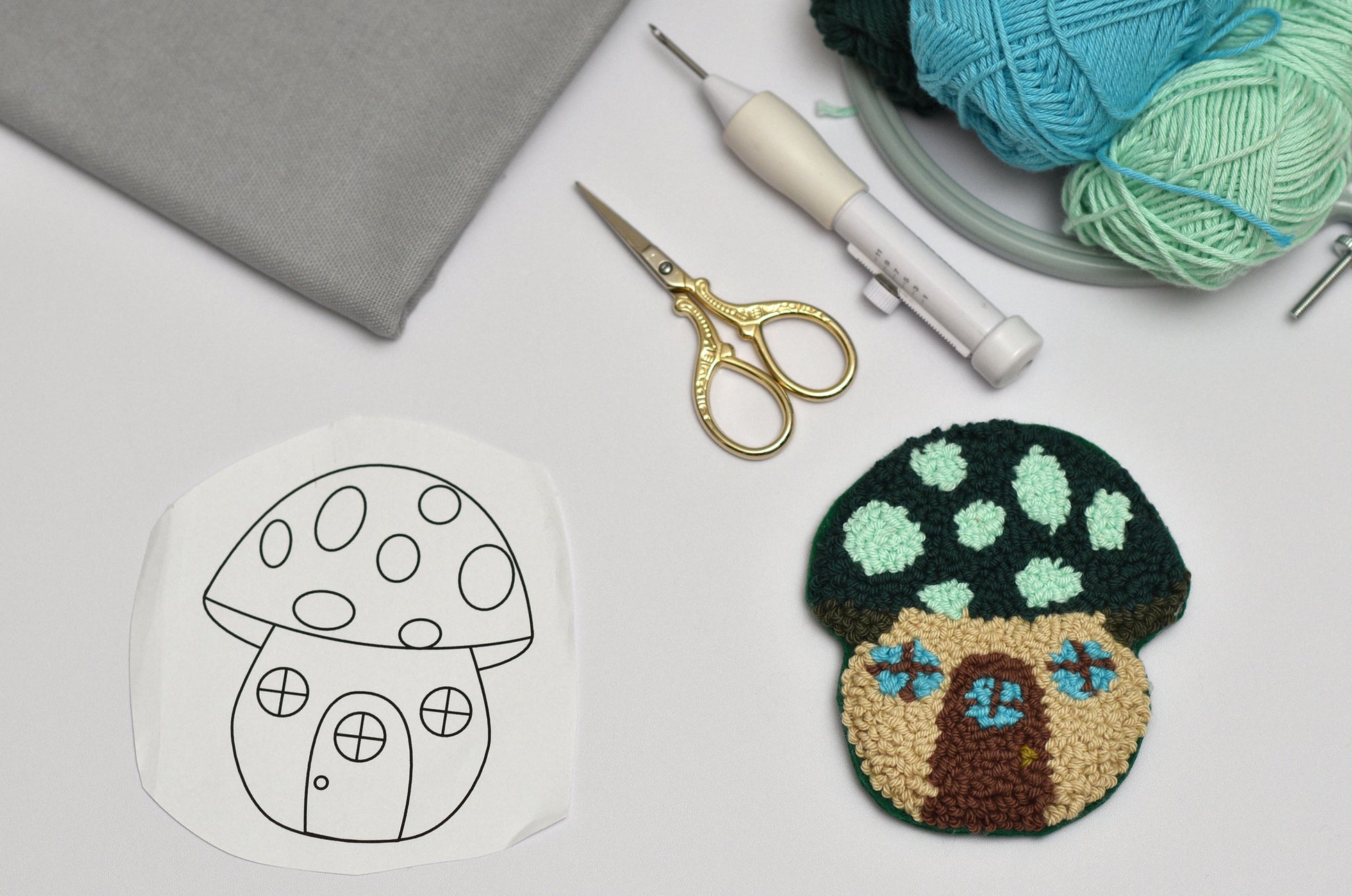 Embroidery hoop with mushroom design, completed mushroom punch needle coaster, scissors, and yarn on a white surface.
