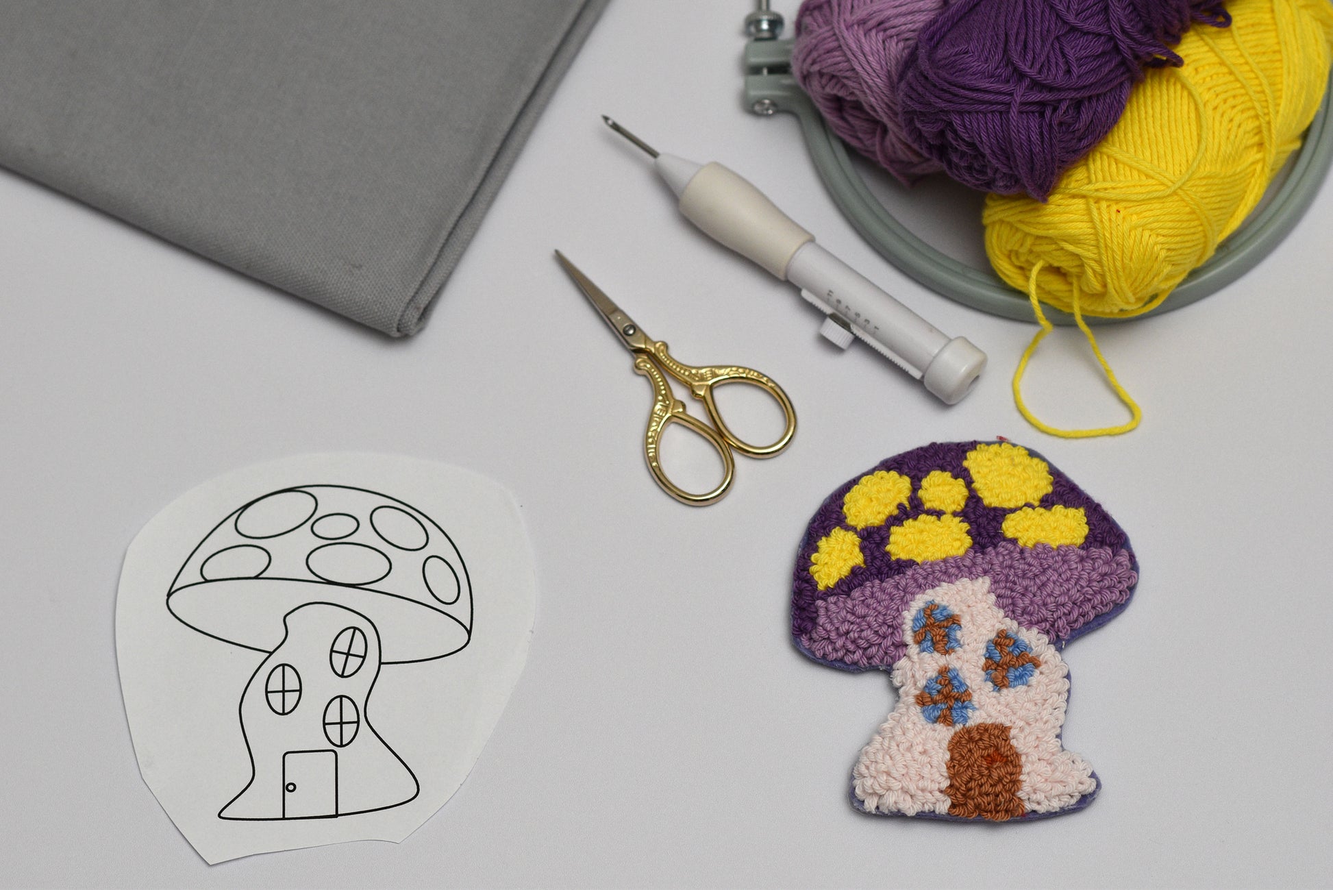 Embroidered mushroom coaster next to yarn, scissors, and a template on a gray surface