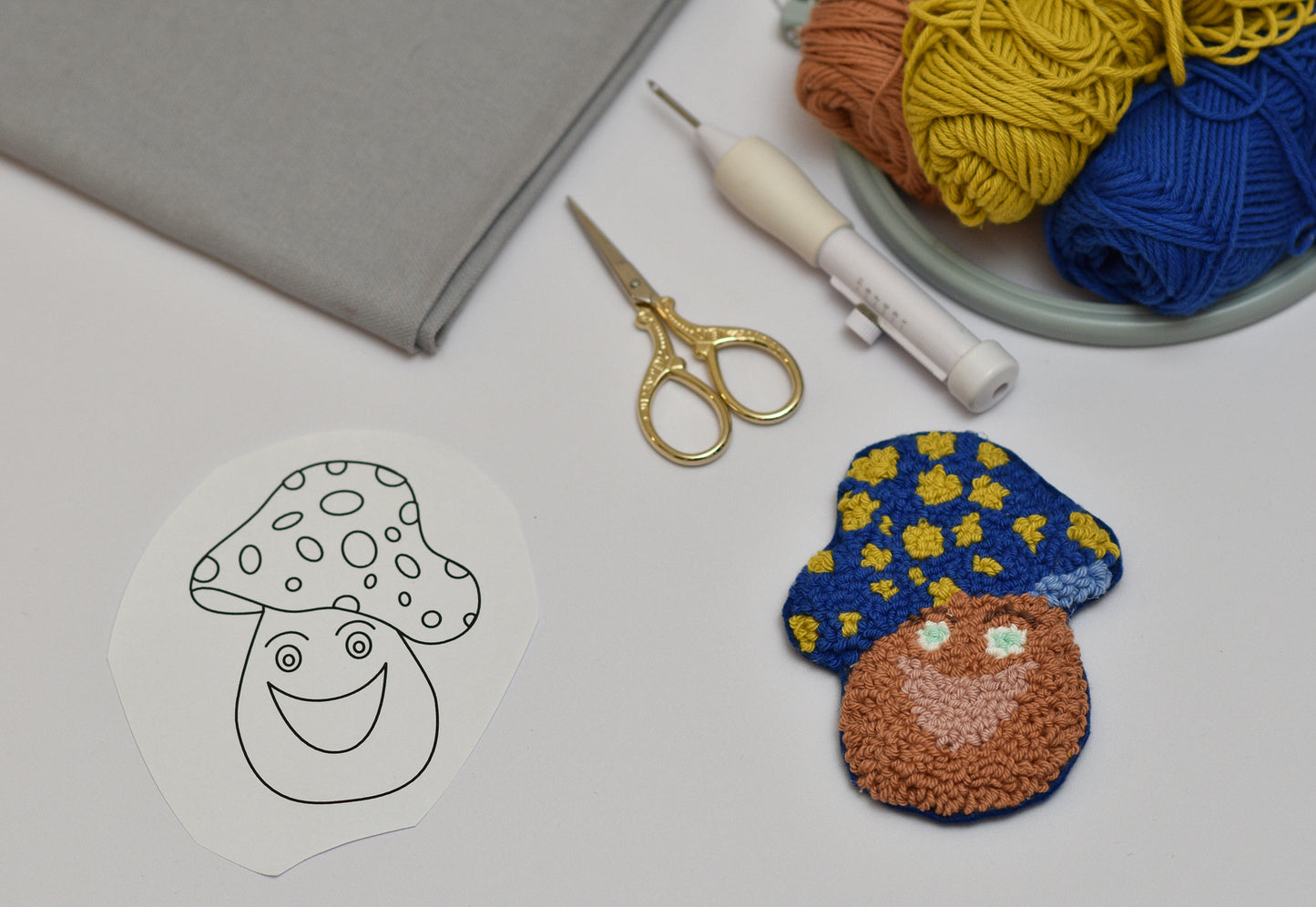 Embroidered mushroom coaster with blue hat and brown body next to a line drawing of a mushroom on a table with yarn and tools.