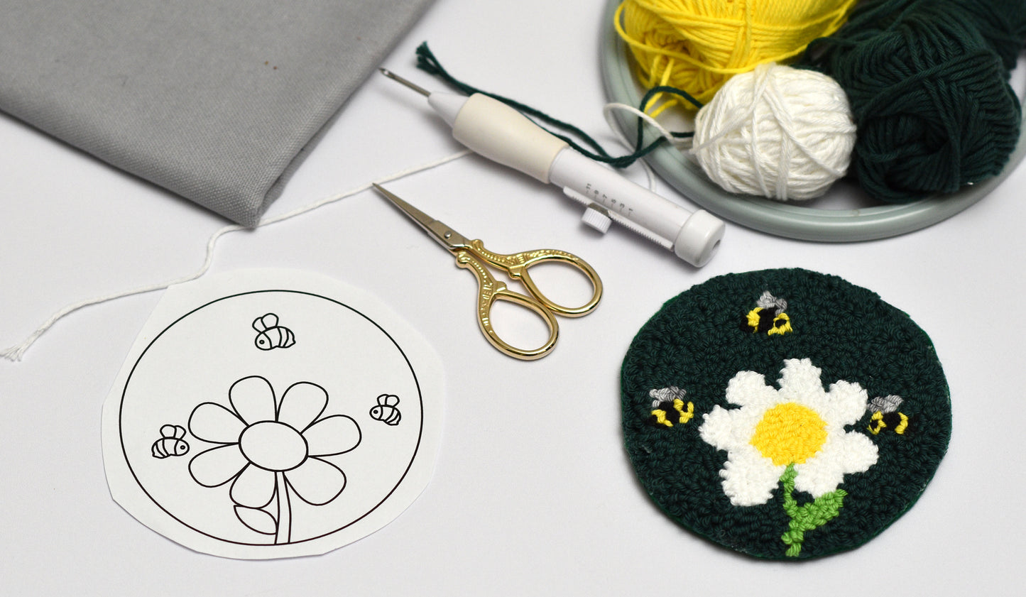 Embroidery hoop with a completed coaster with daisy and bee design, yarn, scissors, and pattern on a white surface.
