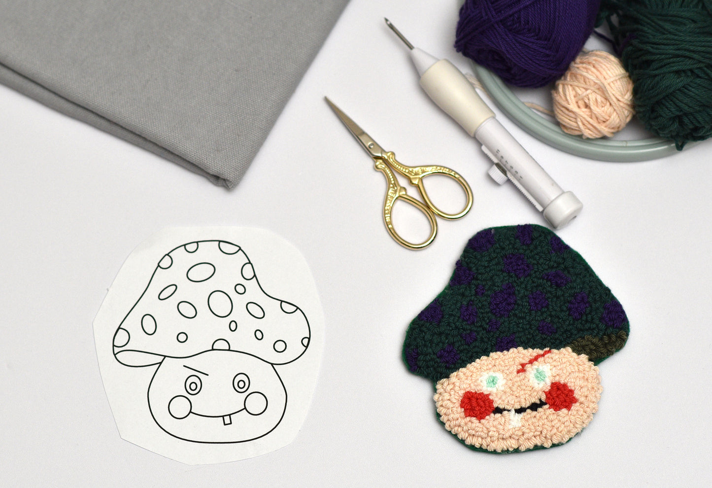 Punch needled coaster with mushroom designed character next to a black and white outline drawing on a white surface with yarn and scissors.