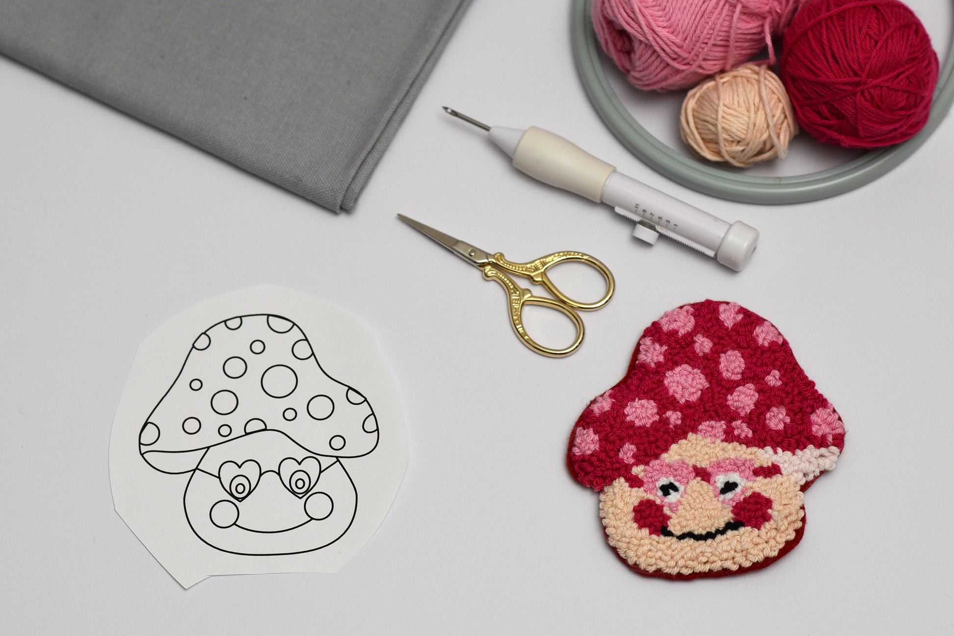 Punch needled coaster with mushroom character head with yarn, scissors, and pattern on a light surface