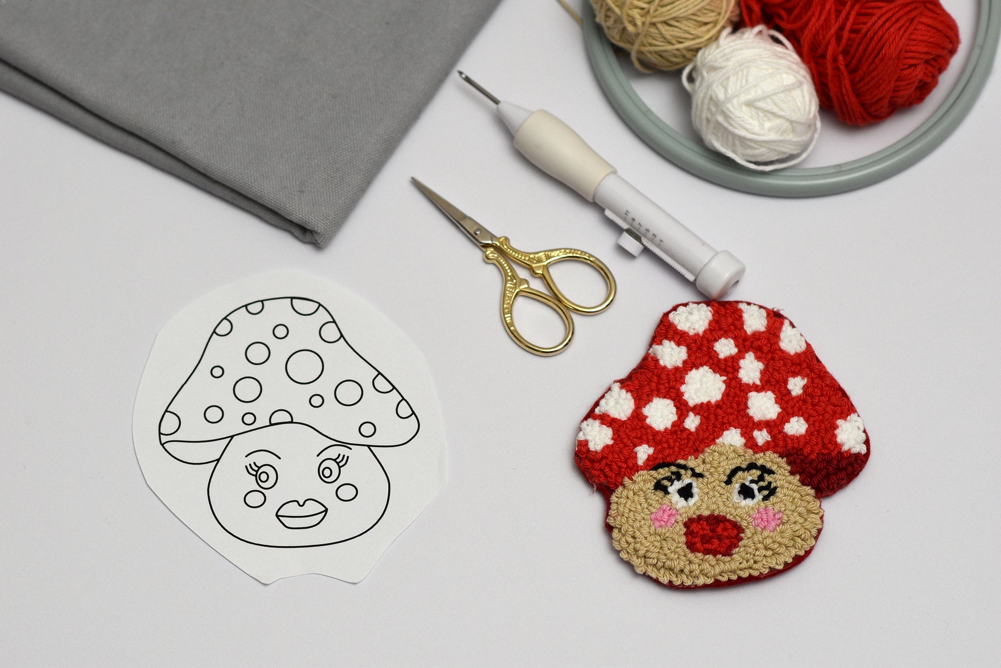Punch needled coaster with mushroom with face next to a drawing of a mushroom, scissors, and yarn on a light surface.