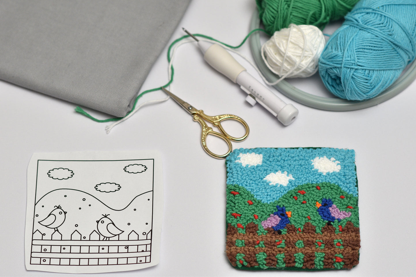 Embroidered coaster with a landscape design next to a coloring sheet and knitting supplies on a table.