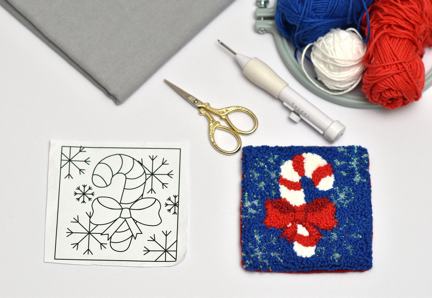 Punch needle square coaster with candy cane and bow design next to a pattern sheet, scissors, and yarn on a white surface.