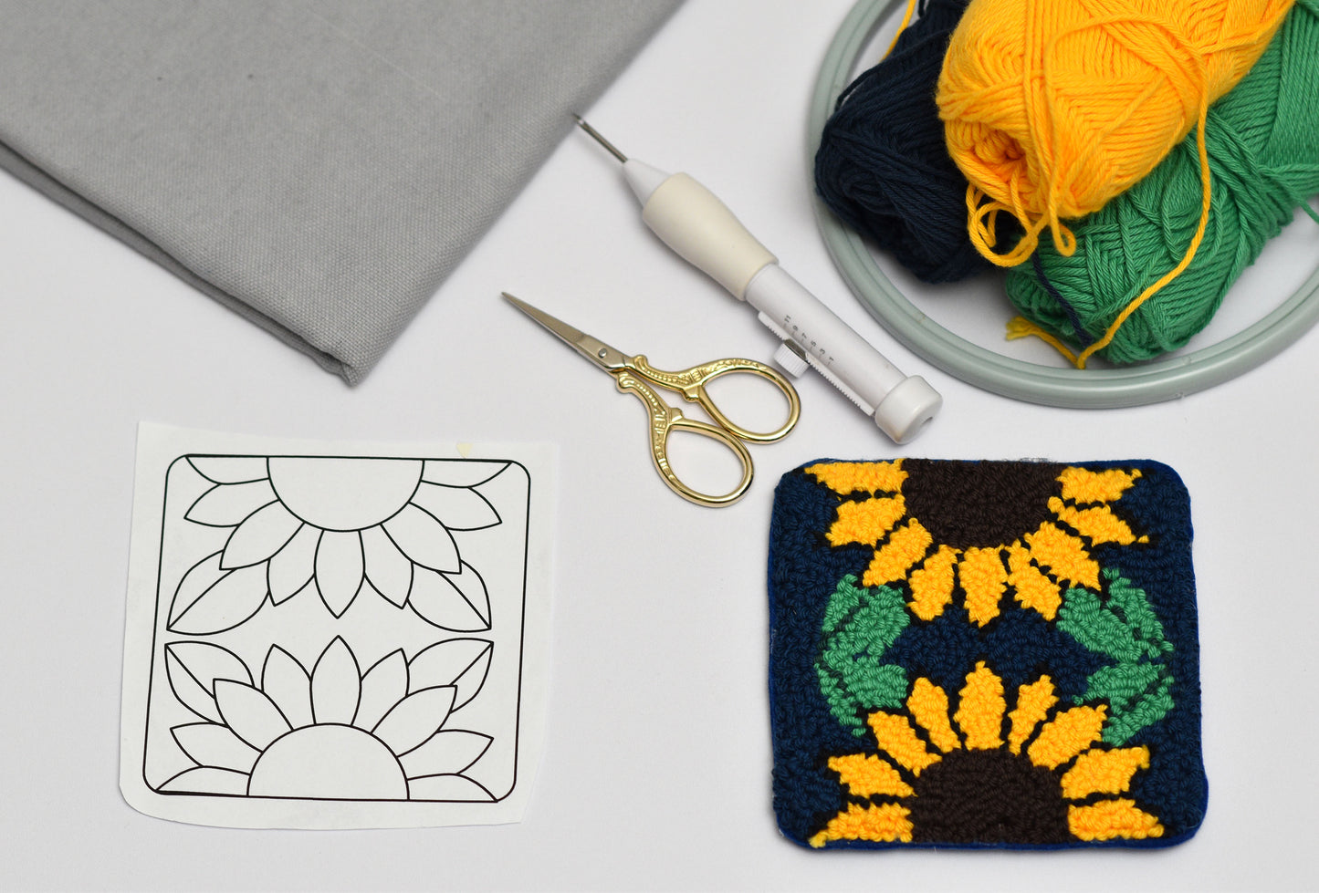 Punch needled sunflower coaster pattern next to a black and white outline, scissors, and yarn on a gray surface.